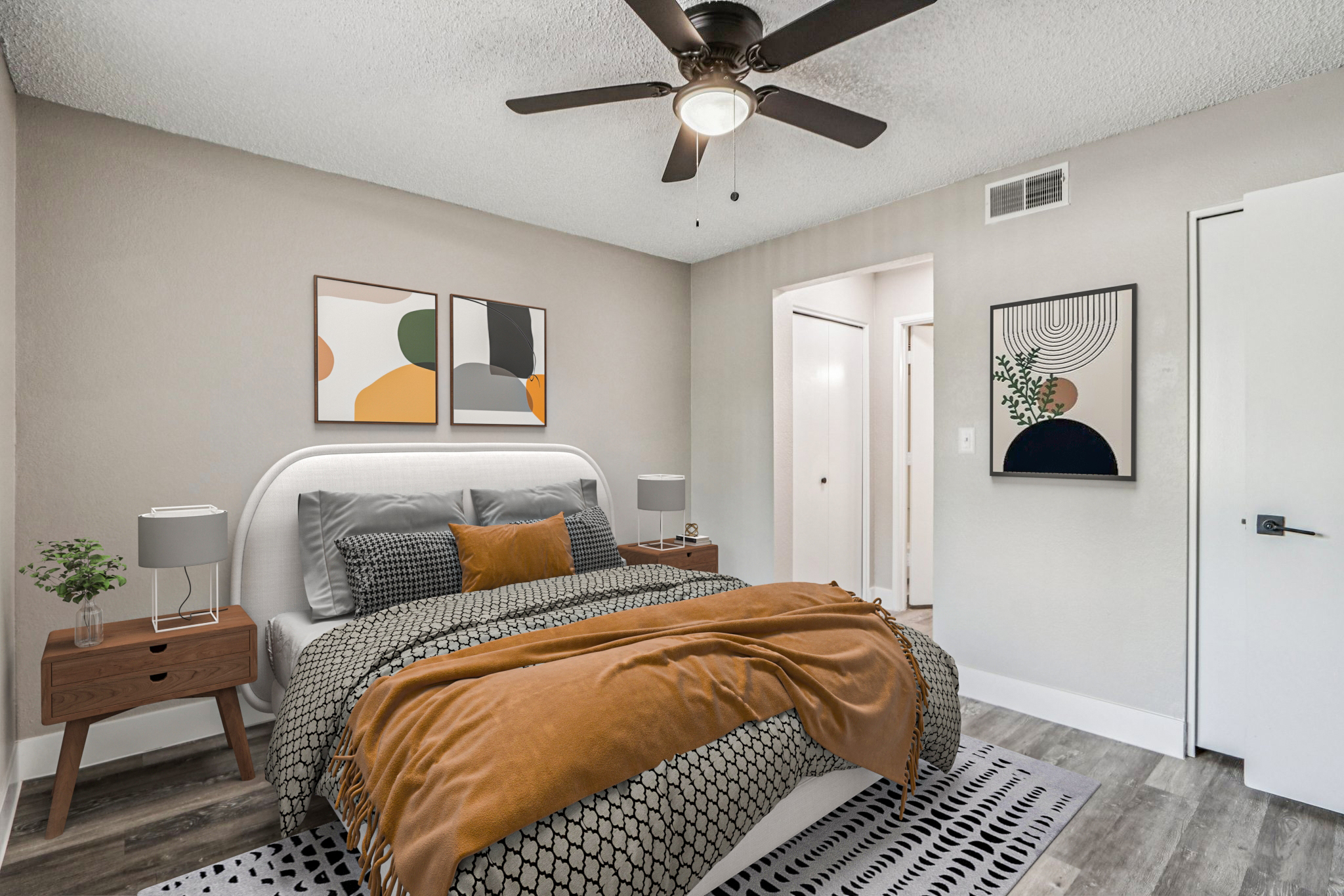 A cozy bedroom featuring a large bed with a gray headboard and orange throw blanket. Two modern abstract paintings hang on the wall. There are wooden nightstands on either side of the bed, with a small plant on one. A ceiling fan is visible above, and a doorway leads to another room. The decor is neutral and inviting.