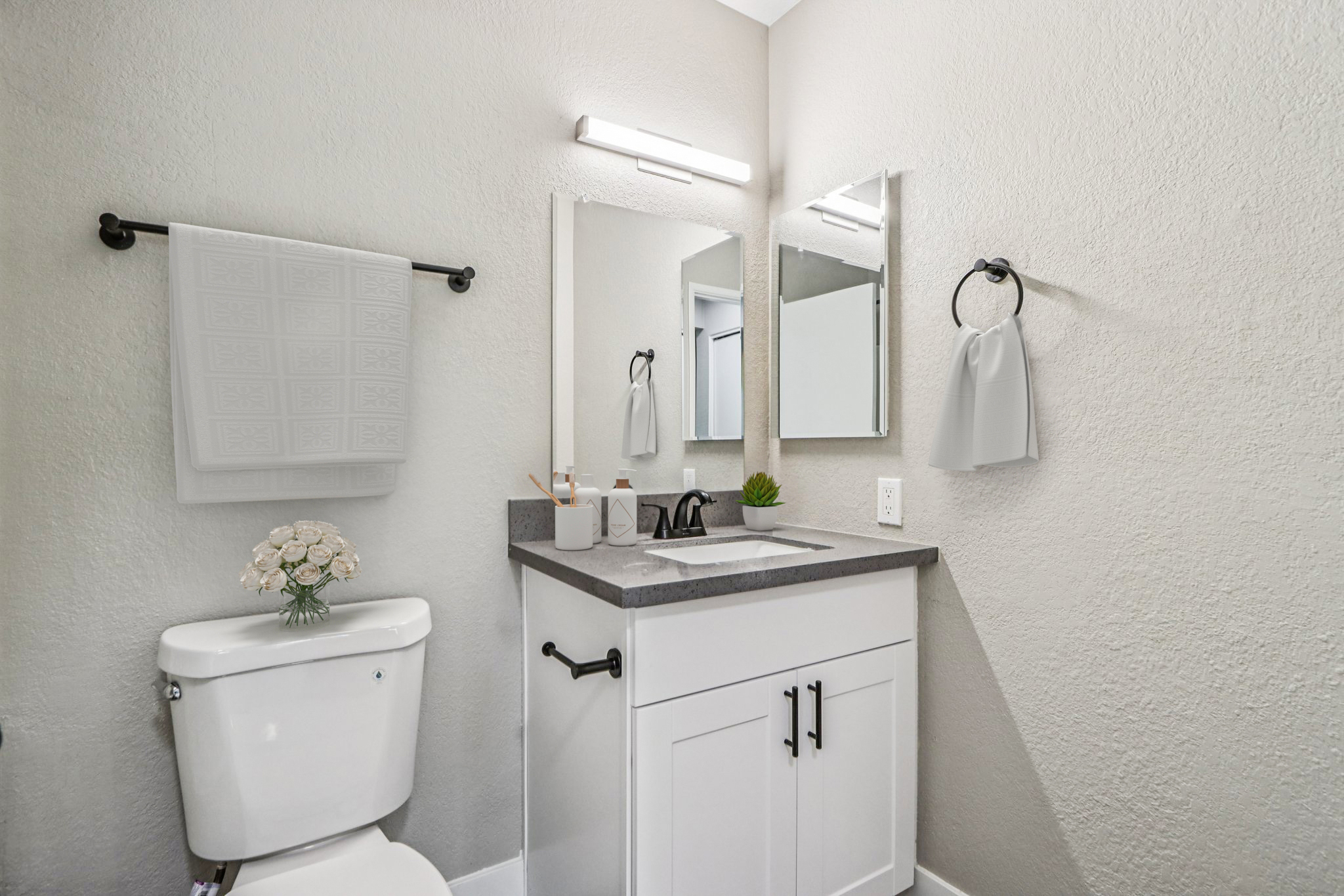 A modern bathroom featuring a white toilet, a sleek vanity with a sink, and a large mirror. The walls are painted a light gray, and there are decorative elements, including a small plant and a vase with flowers. A towel hangs on a rack, adding a touch of warmth to the space.