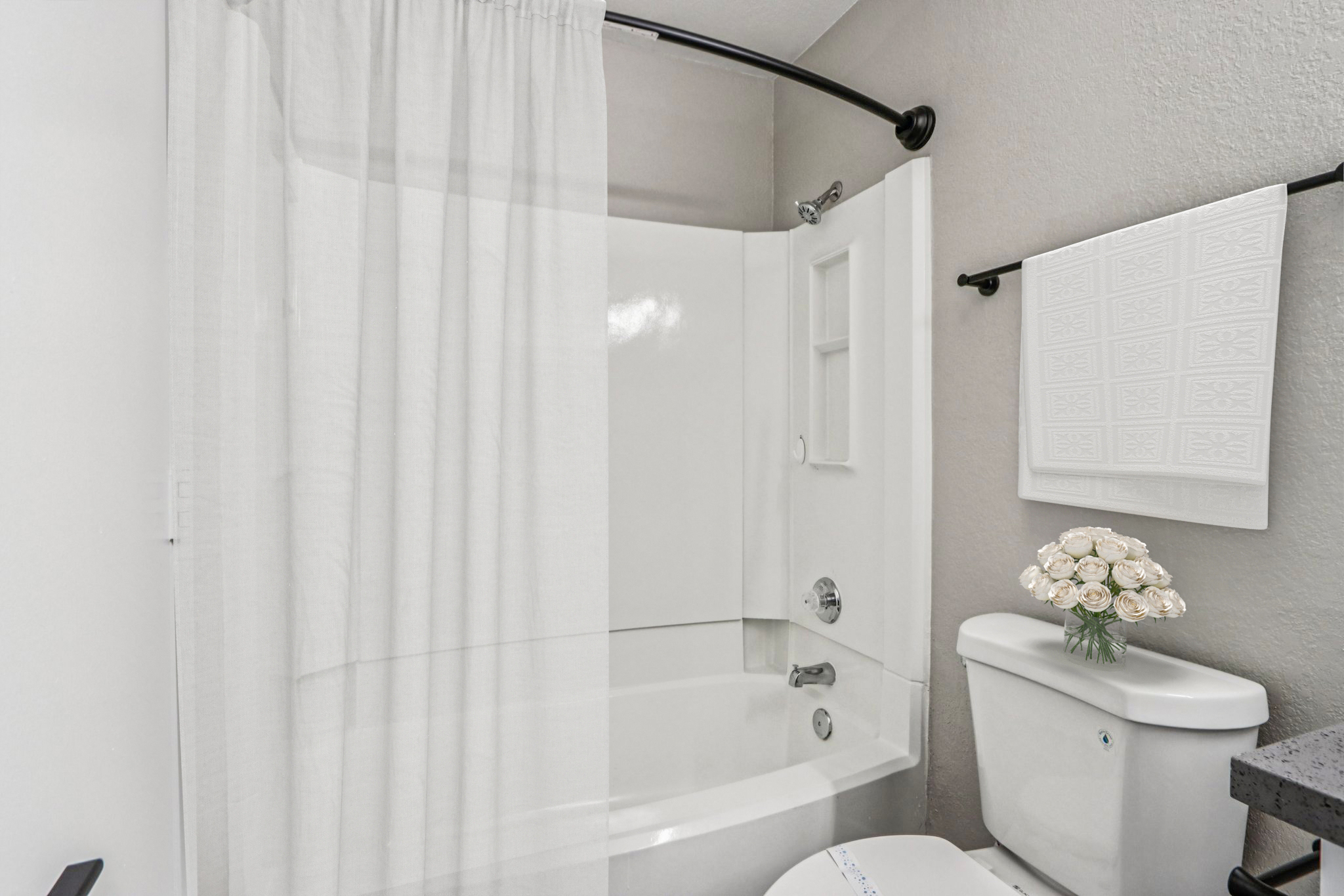 A clean, modern bathroom featuring a bathtub with a white shower curtain, a toilet, and a small vase of white flowers on a countertop. The walls are painted in a light color, and a decorative towel is hanging on a bar. The overall ambiance is minimalist and fresh.