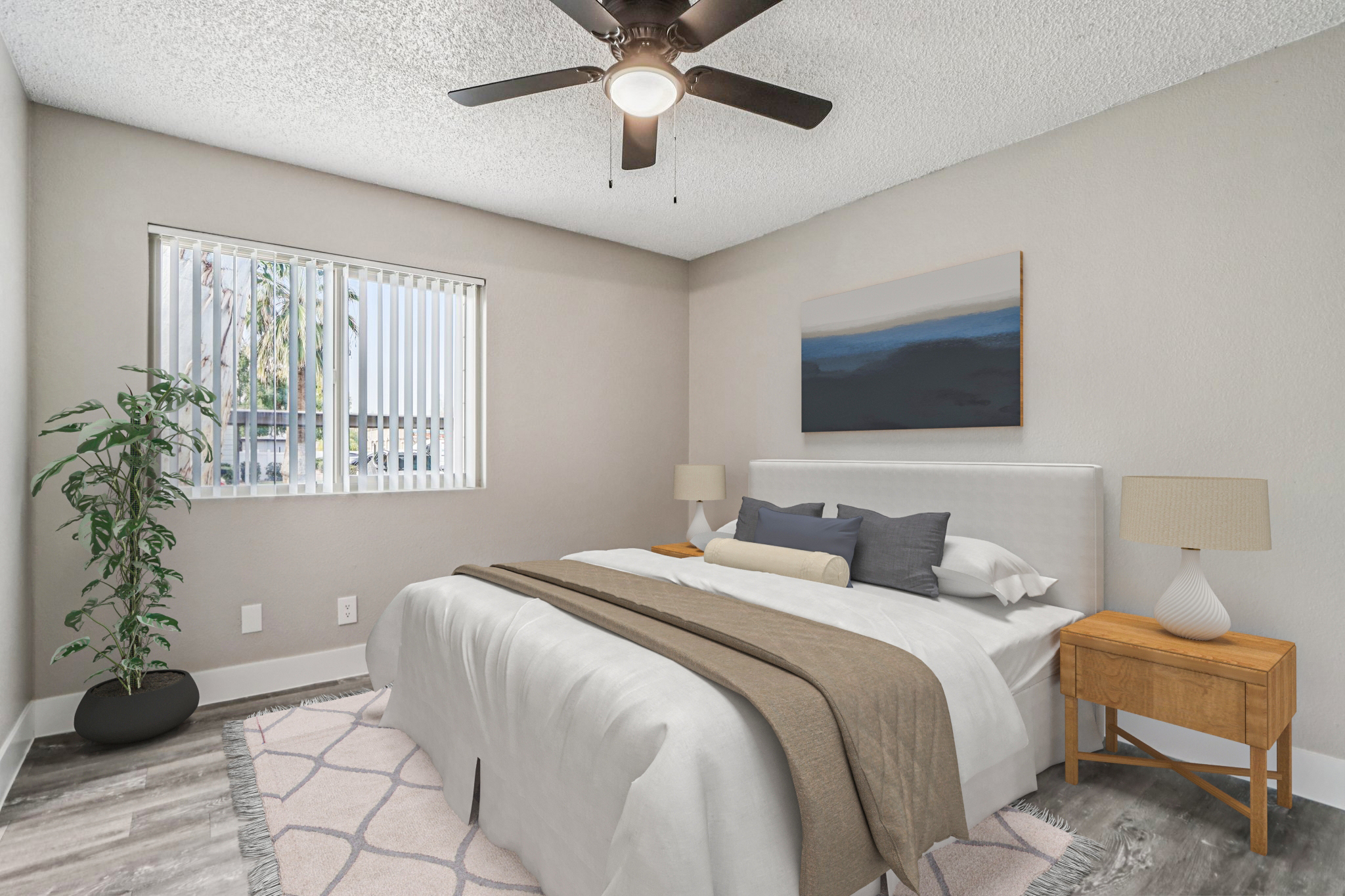 A cozy bedroom featuring a white bed with gray and beige linens, two bedside lamps on wooden nightstands, and a ceiling fan. A large window with vertical blinds allows natural light, while a potted plant adds a touch of greenery. A modern artwork hangs on the wall above the bed, complementing the neutral color scheme.