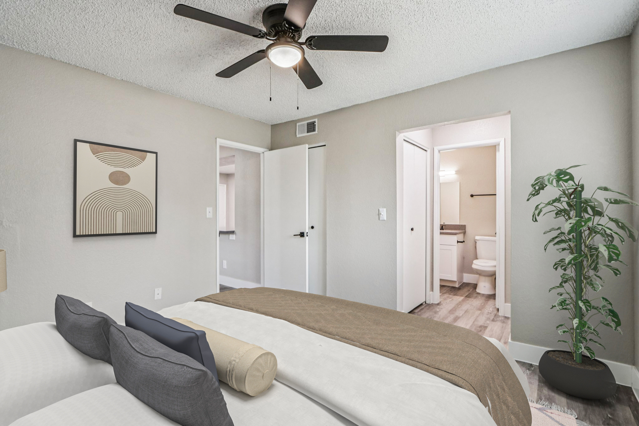 A well-decorated bedroom featuring a bed with neutral bedding, a ceiling fan, and a decorative plant. The room has light-colored walls and a piece of wall art, with a view into a bathroom and a closet visible through open doors in the background.