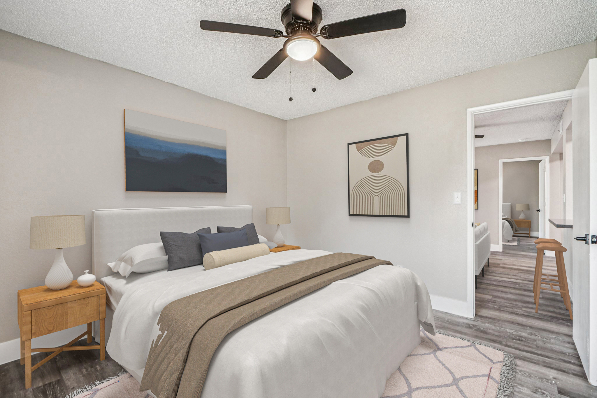A cozy bedroom featuring a neatly made bed with white linens and a brown throw blanket. There are two wooden side tables with lamps, and the walls are painted in a soft beige. A piece of abstract art hangs above the bed, and a doorway leads to another part of the home, visible in the background.