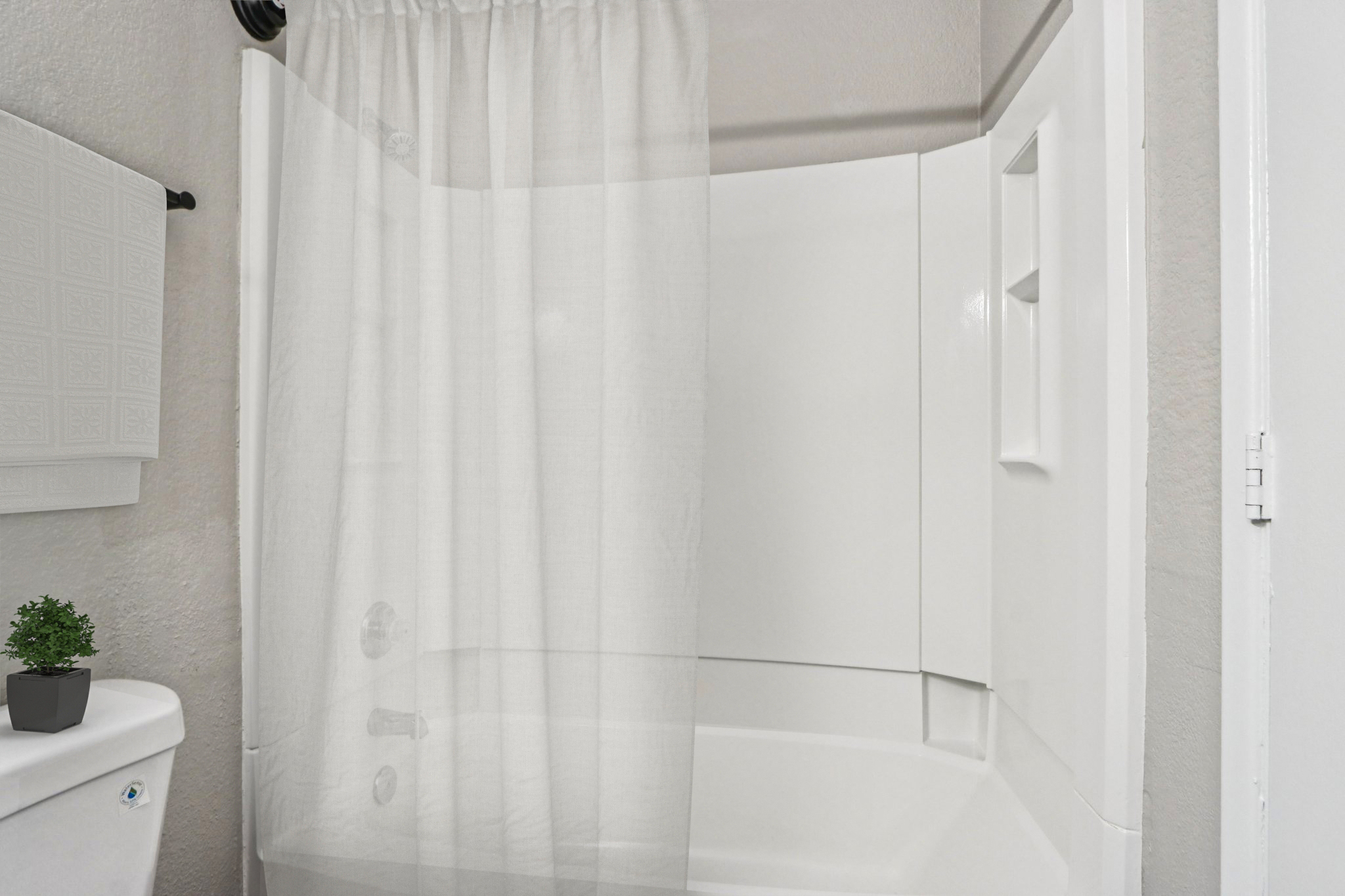 A bathroom scene featuring a white bathtub with a tall, sheer shower curtain partially closed. A small green plant is placed on a white toilet next to the shower area. The walls are light-colored, and there's a simple shelf or wall piece visible. The overall atmosphere is clean and minimalist.
