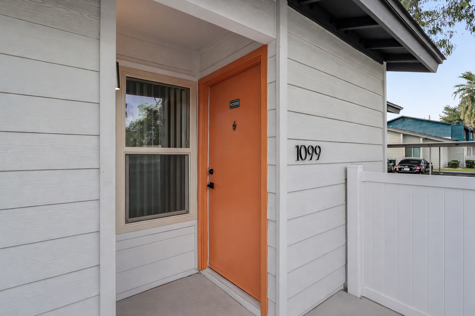 Front entrance of a house featuring an orange door, light-colored siding, and a clear house number "1099" displayed on the exterior wall. A white picket fence runs along the front, and there are trees and additional houses visible in the background.