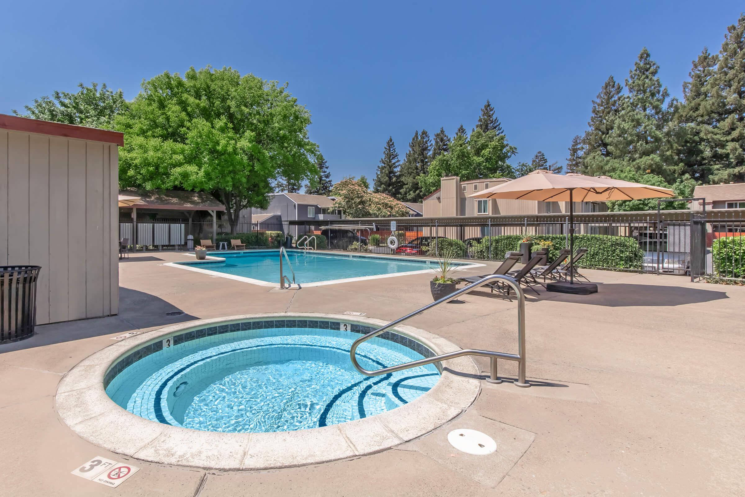 A sunny outdoor area featuring a swimming pool and a hot tub. Surrounding the pool are lounge chairs and umbrellas, with trees visible in the background. The space is well-maintained and includes a fence for safety, creating a relaxing environment for leisure and recreation.