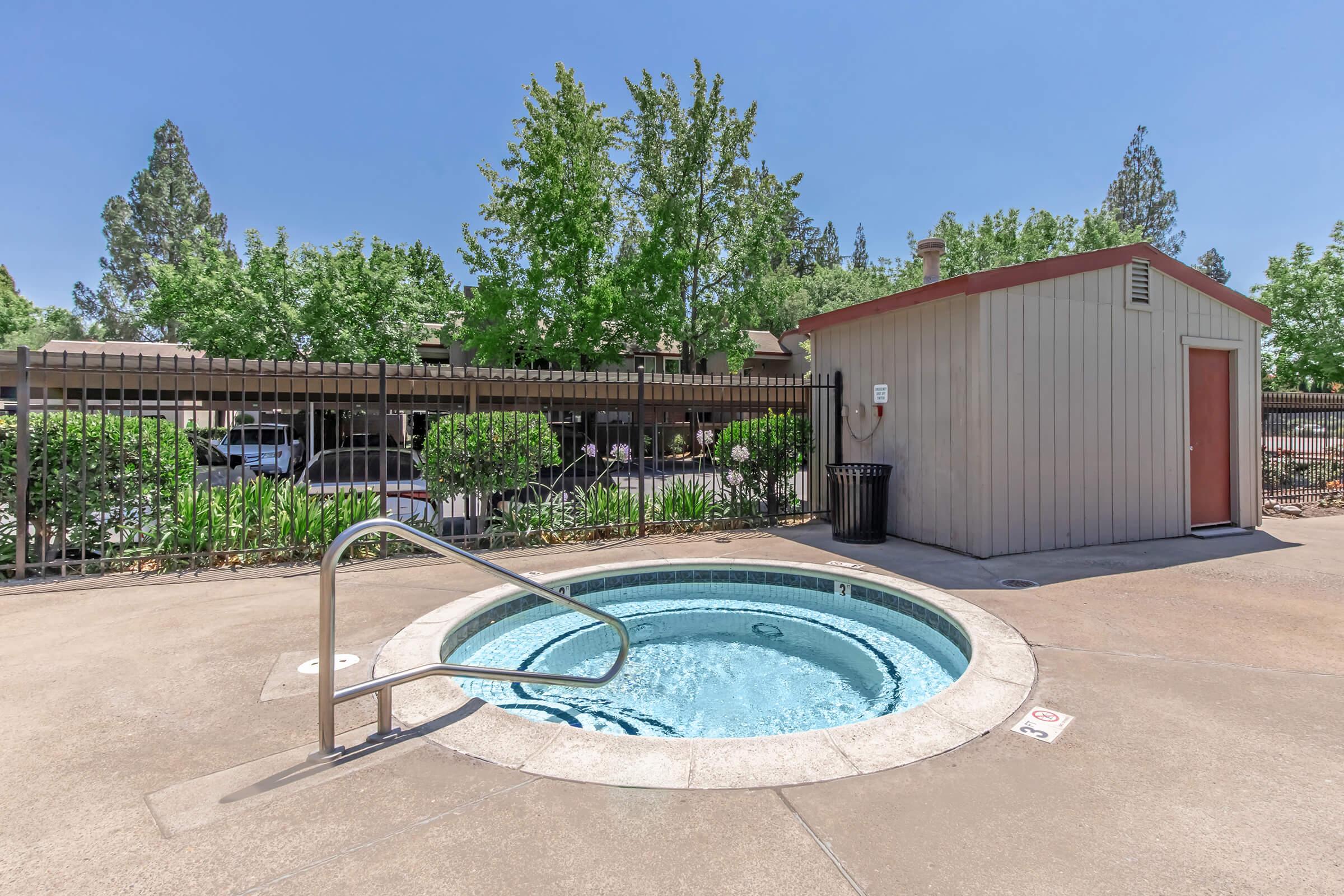 A small outdoor hot tub surrounded by concrete. In the background, there are trees and a building with a fence. A nearby shed has a door, and a trash can is visible. The sky is clear and blue, indicating sunny weather.