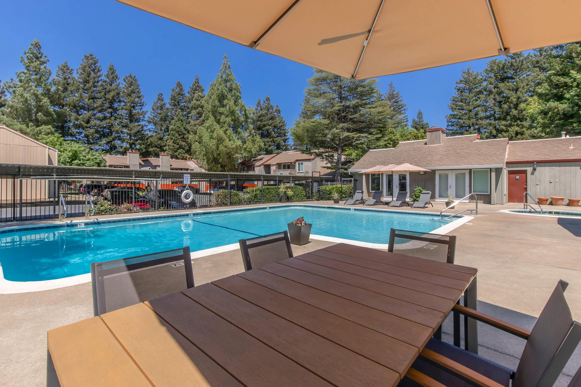 A spacious outdoor area featuring a swimming pool surrounded by tall trees. In the foreground, there is a wooden dining table with chairs shaded by a large umbrella. The pool area includes lounge chairs and is enclosed by a fence, with residential buildings visible in the background under a clear blue sky.