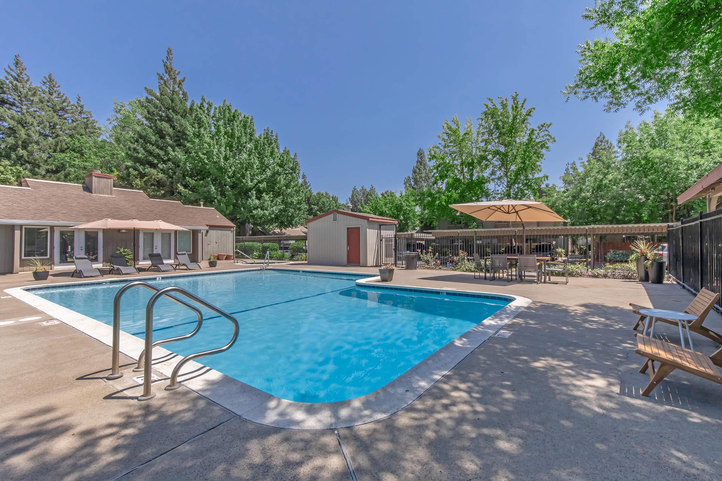 A clear blue swimming pool surrounded by trees and lounge chairs. A large umbrella provides shade near the poolside, with a spacious area featuring tables and chairs in the background. The setting is bright and inviting, ideal for relaxation and leisure.