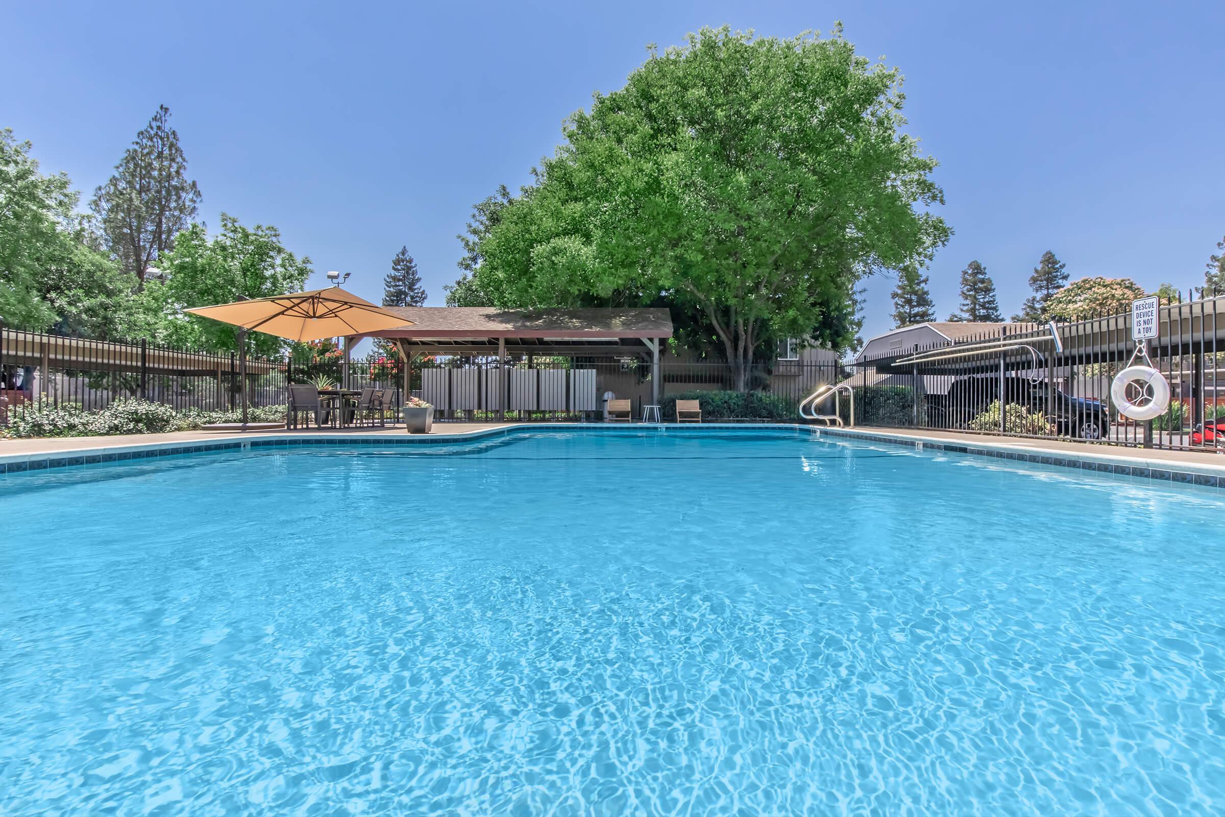 A brightly lit swimming pool surrounded by a fence, featuring a shaded area with an umbrella and lounge chairs. Lush green trees are visible in the background under a clear blue sky. The water appears inviting and clean, making it an ideal spot for relaxation and leisure.