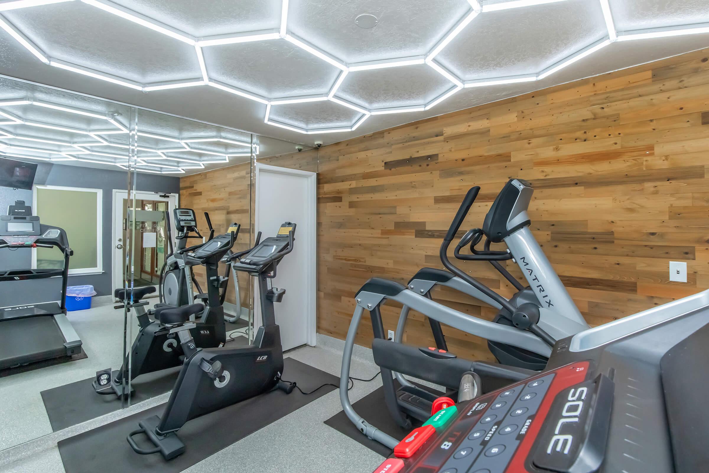 A modern gym interior featuring exercise equipment like treadmills, elliptical machines, and stationary bikes. The space has a unique hexagonal ceiling design with LED lighting and wooden walls, creating a contemporary and inviting atmosphere for workouts.