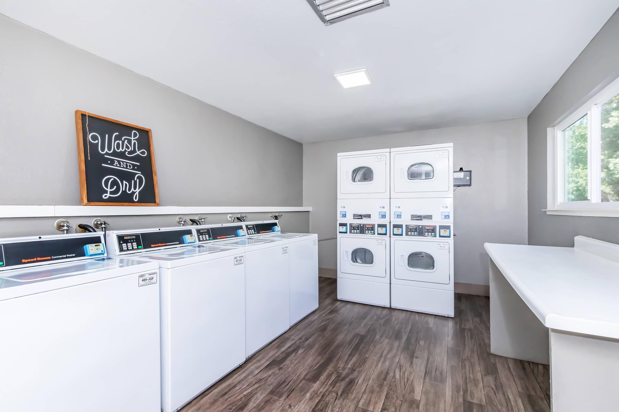 A clean laundry room with white washing machines and dryers lined up against a wall. A sign reading "Wash and Dry" is displayed on the wall. The room features light-colored walls, a large window allowing natural light, and a folding counter. The floor is wooden, adding to the modern aesthetic.