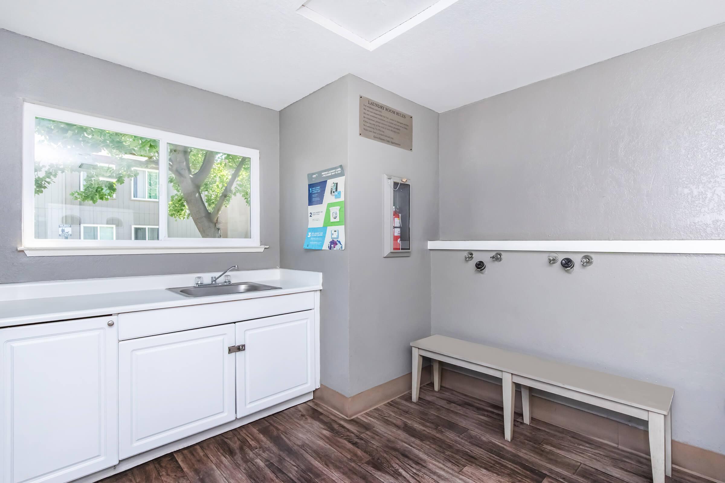 A clean and modern laundry room featuring a white cabinet and sink, a bench against the wall, and a window that lets in natural light. informational posters are displayed on the wall next to a fire extinguisher, with gray walls and wood-like flooring creating a contemporary atmosphere.
