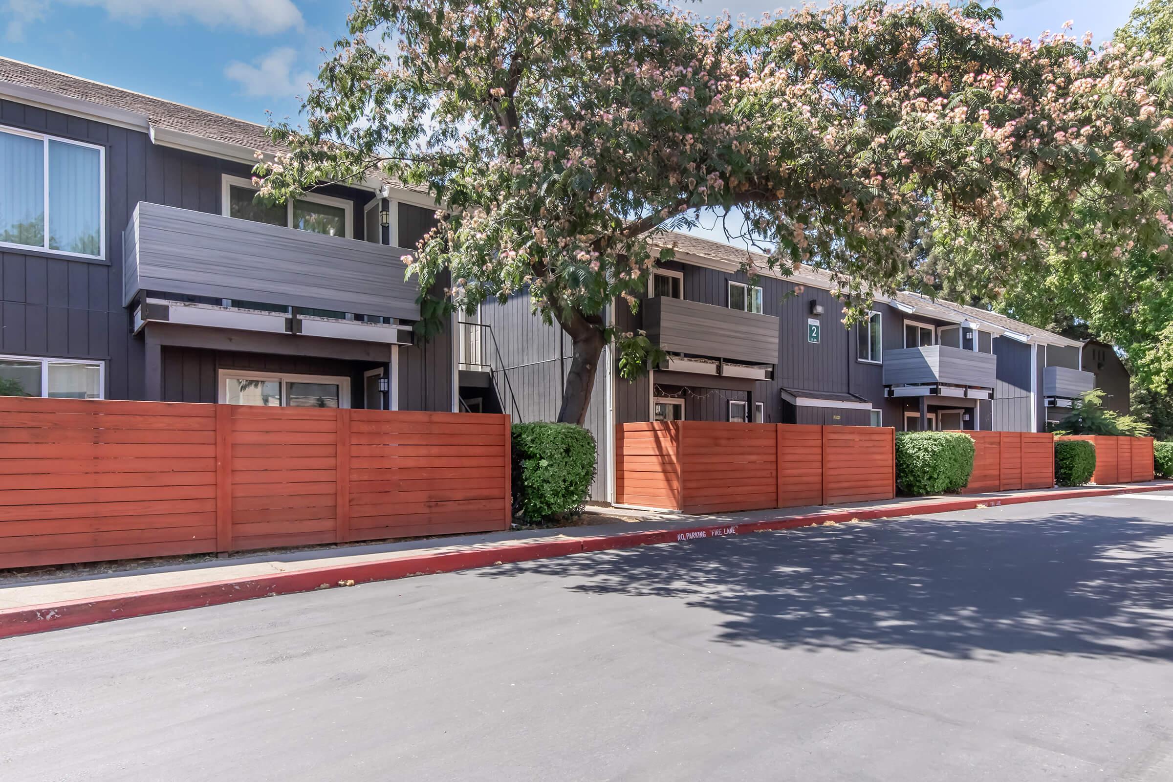 A row of modern apartment buildings with gray siding, featuring balconies and large windows. The area is lined with freshly painted wooden fences and neatly trimmed bushes. In the foreground, a smooth, empty roadway provides access to the complex, with a clear blue sky above and trees dotting the scene.