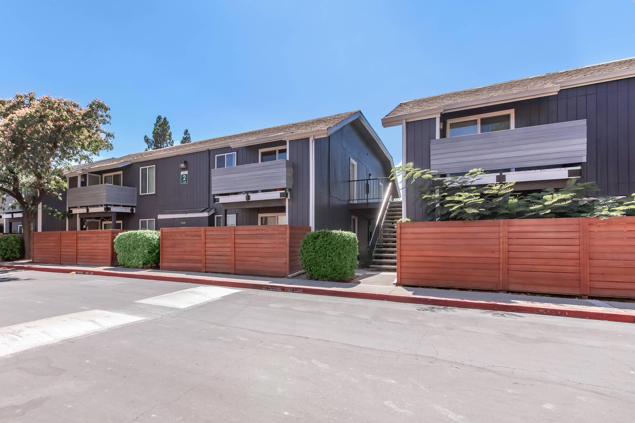 Two-story apartment buildings with dark exteriors and wooden balconies are seen along a paved path. Neatly trimmed bushes and wooden fences provide privacy. A clear blue sky is visible above, indicating a bright, sunny day. The scene conveys a peaceful residential environment.