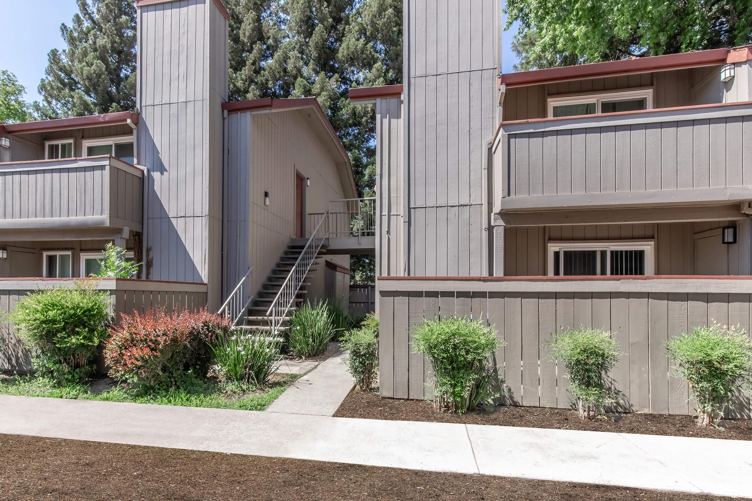 A view of a multi-story apartment building with a light gray exterior, surrounded by greenery. The building features balconies, a stairway, and a walkway leading through a landscaped area with shrubs and trees. Clear skies indicate a sunny day.