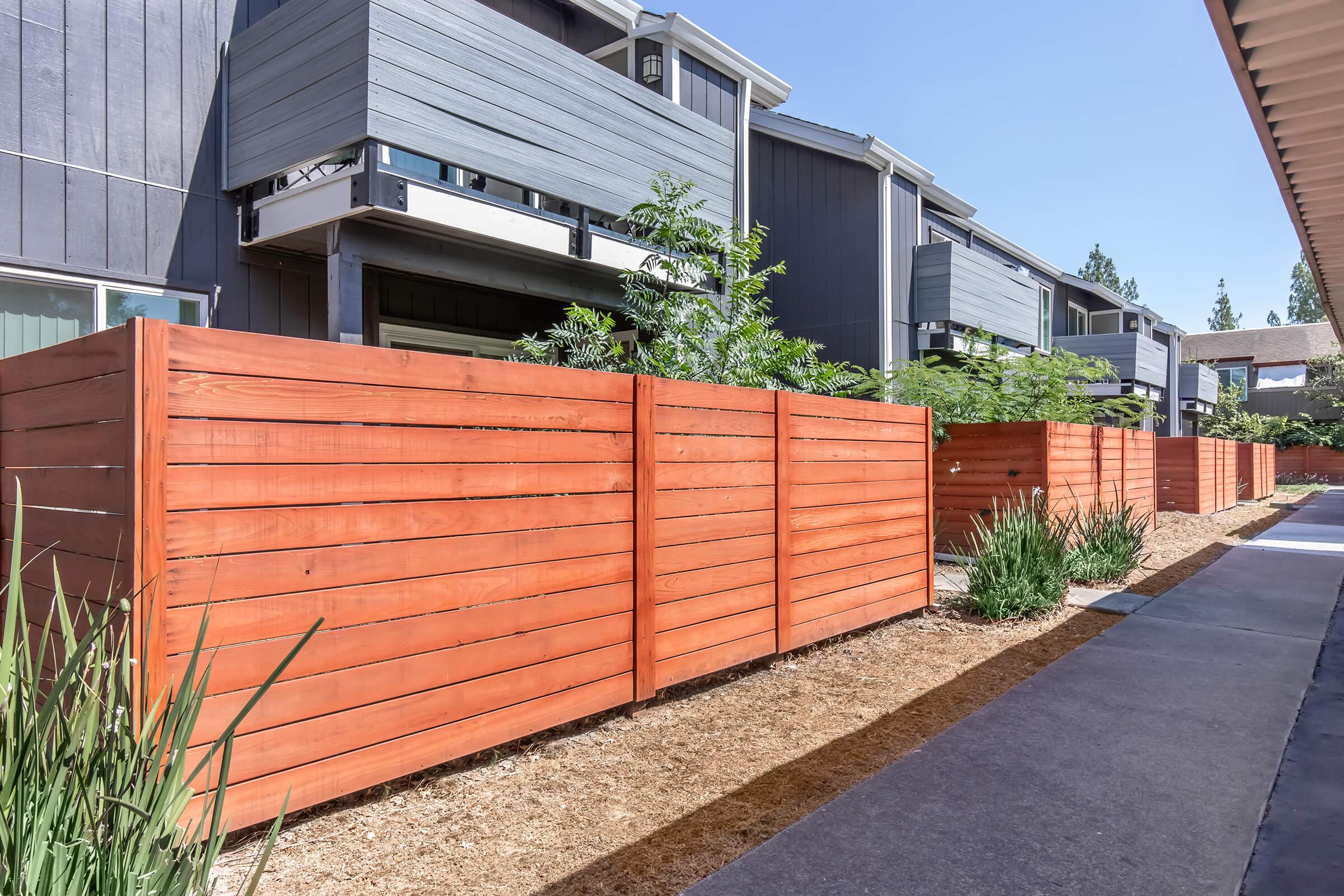 A pathway lined with modern wooden fences and low greenery, leading up to multi-story buildings. The scene features a clean, landscaped area with gravel and plants, showcasing a contemporary residential environment. Sunlight casts gentle shadows on the pavement.