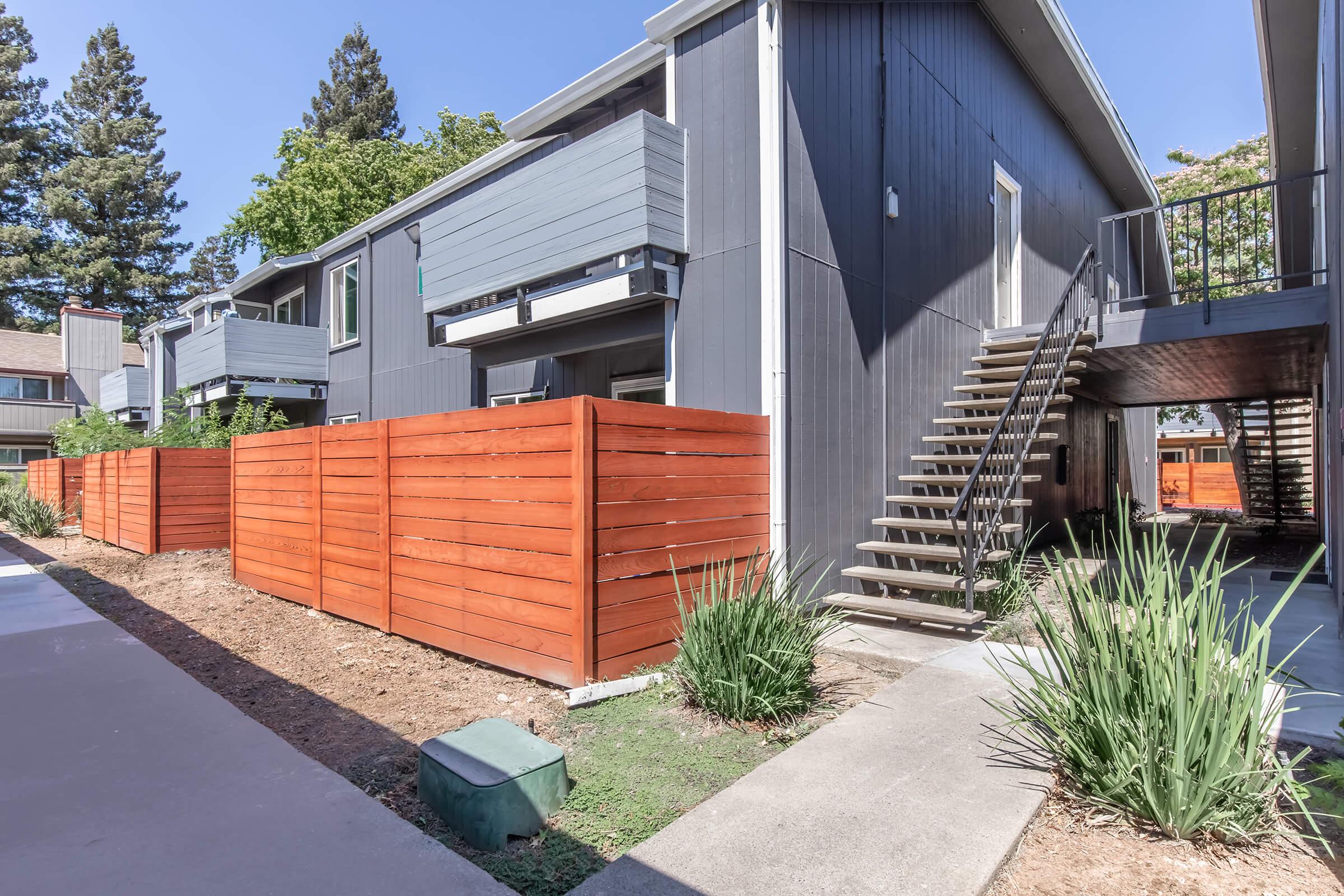 A modern apartment complex featuring gray exterior walls, wooden fences, and a staircase. The scene includes landscaped greenery, with steps leading to the upper level of the building, under a clear blue sky.