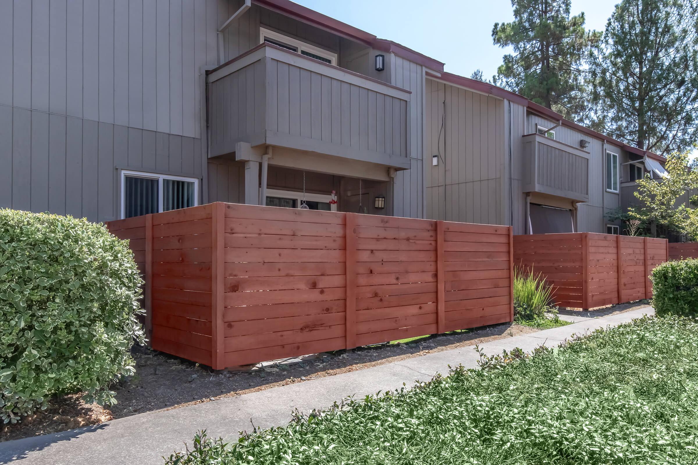 A row of wooden fences surrounds a landscaped area in front of beige apartment buildings. The fences are painted in a reddish-brown color, providing privacy for the outdoor spaces. The scene is bright, with clear blue skies and greenery, including shrubs and grass, lining the pathway.