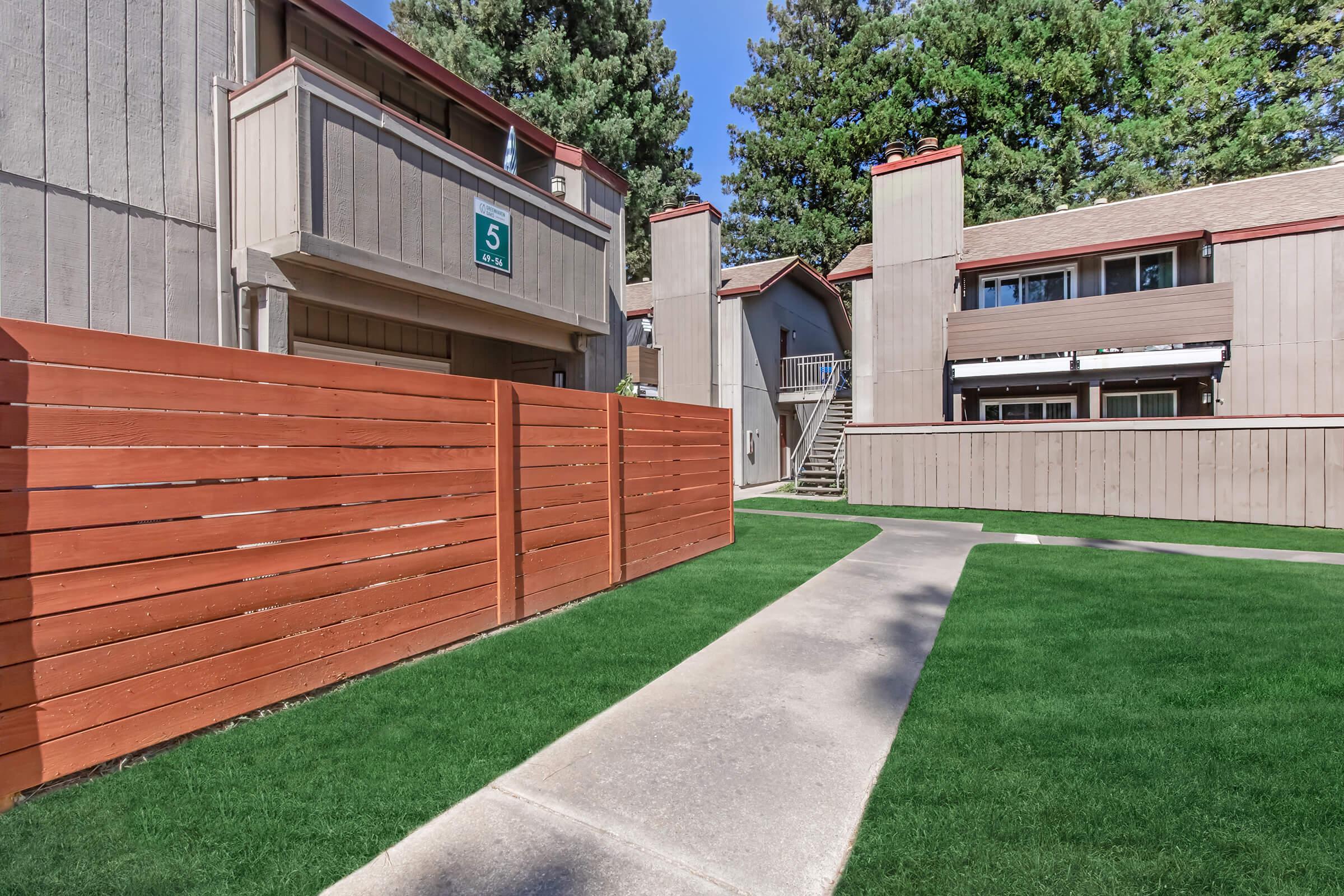 A landscaped pathway leads through a well-maintained residential complex, featuring two-story buildings with balconies. The area is surrounded by wooden fences and lush green grass, with tall trees providing shade in the background. The setting is bright and inviting under a clear blue sky.