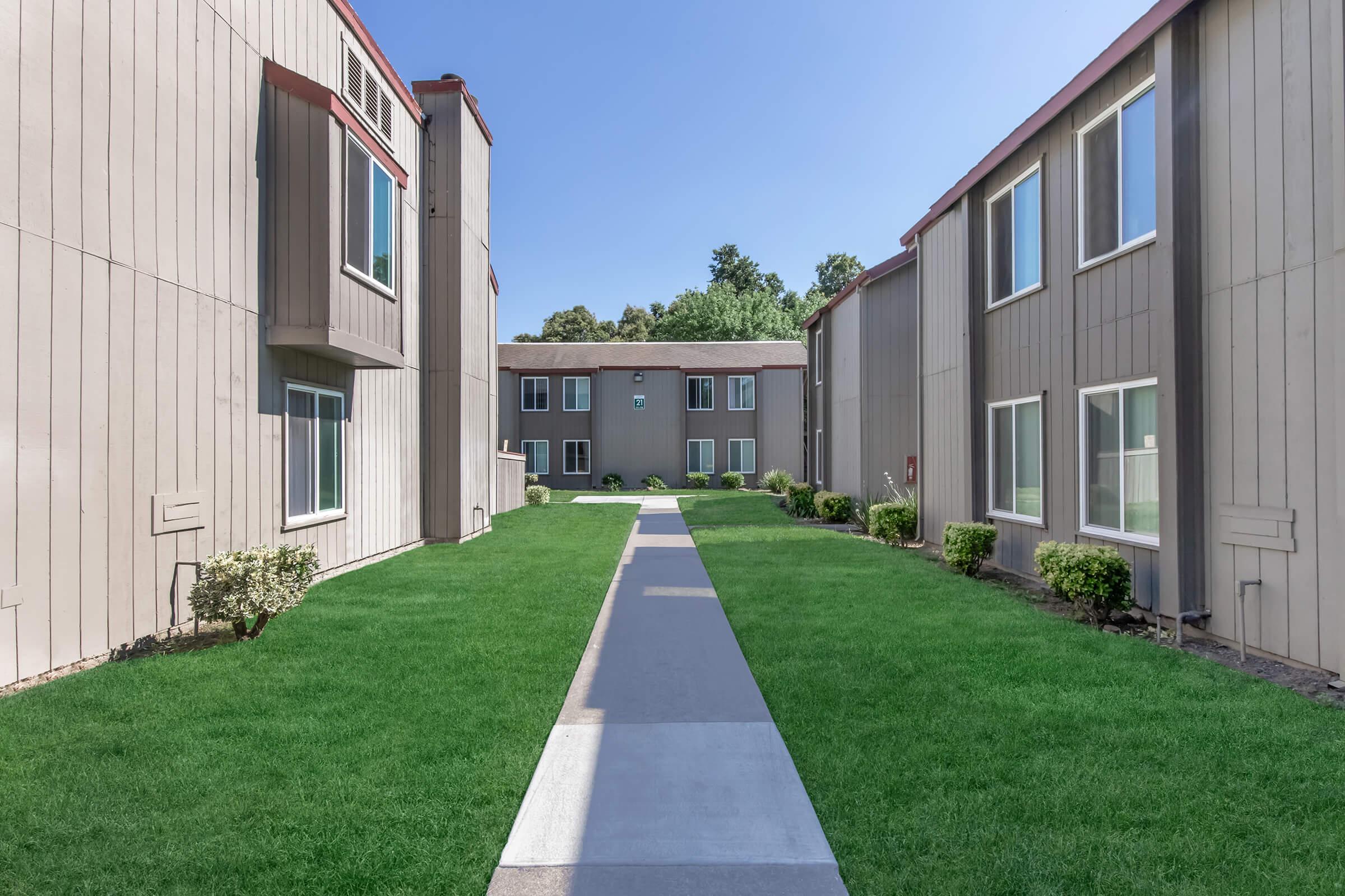 A well-maintained outdoor pathway flanked by two rows of modern apartment buildings, featuring green grass and minimalist landscaping under a clear blue sky. The pathway leads to a central building in the background, creating a welcoming and spacious residential atmosphere.