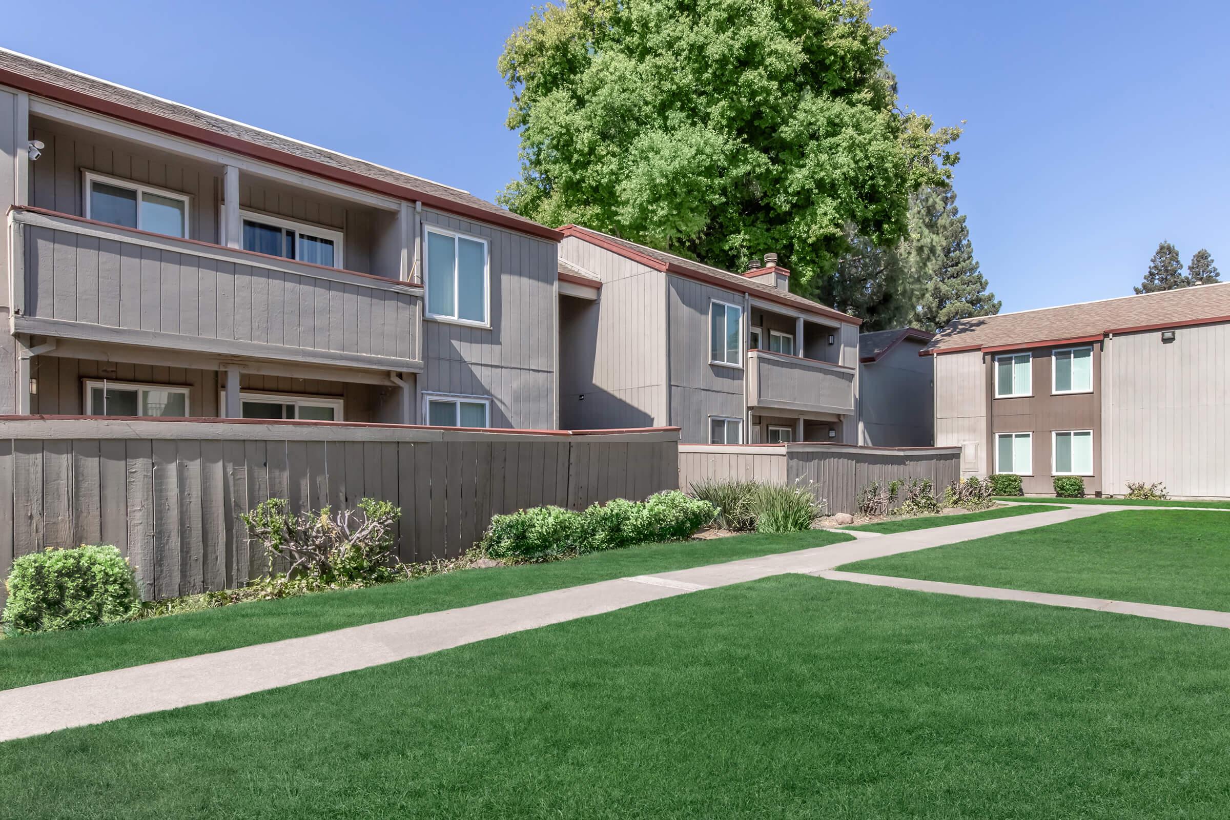 View of a residential apartment complex featuring two-story buildings with gray siding. Lush green grass and neatly landscaped bushes surround the pathways leading to the entrances. A large tree is visible in the background under a clear blue sky.