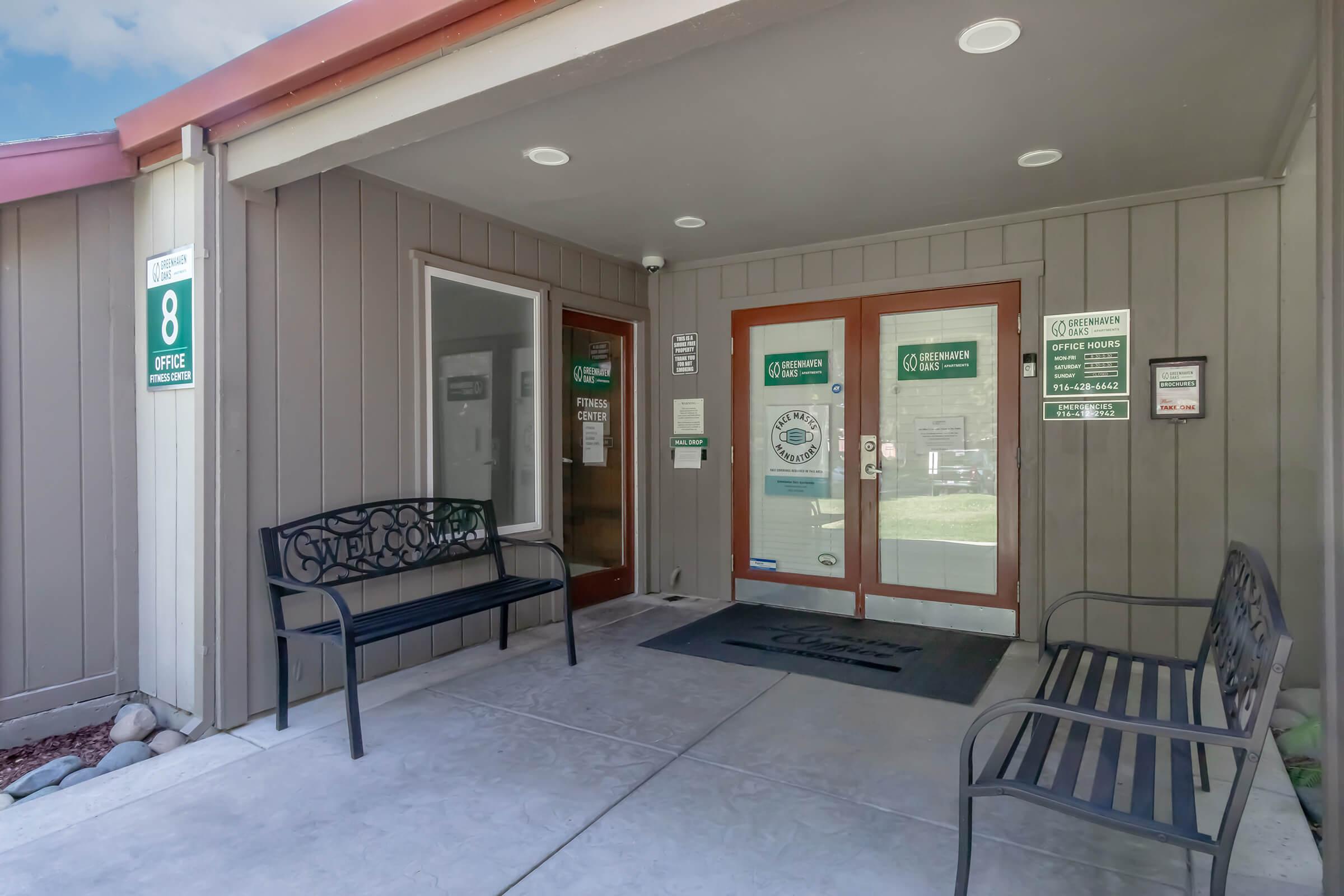 Exterior view of an office entrance featuring double glass doors with signage. Flanking the entrance are two black benches, and a welcome mat is placed in front. The building has a modern design with a light gray exterior and a red roof. Signs indicate office hours and contact information.