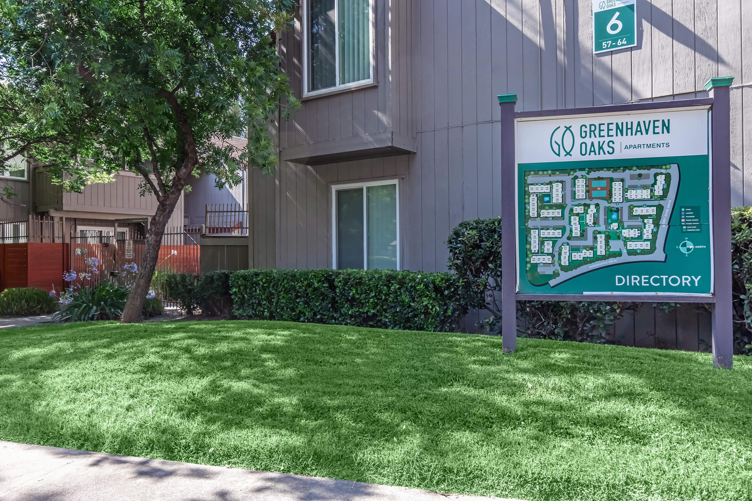 A sign for Greenhaven Oaks Apartments displays a directory map of the complex, set against a background of green grass and shrubbery, with a building visible in the background. The sign is located near the entrance of the property, providing information to residents and visitors.