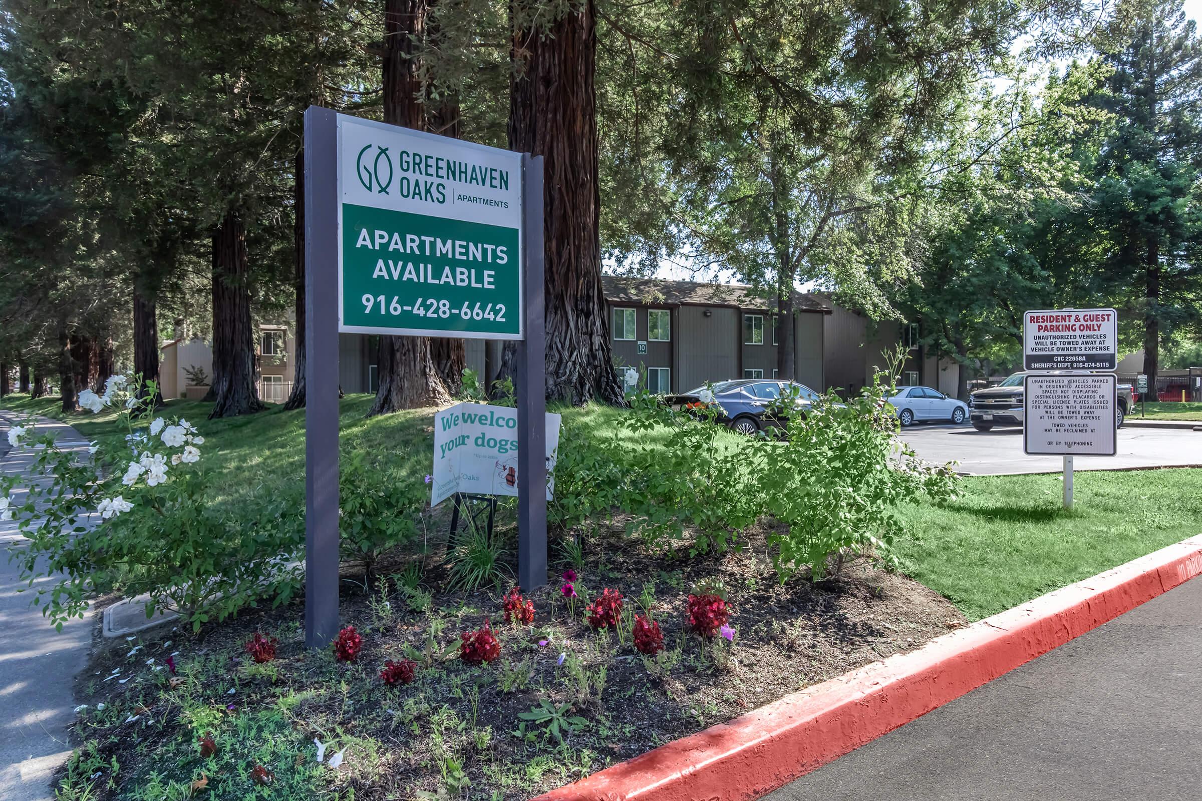 Sign for Greenhaven Oaks Apartments featuring the text "Apartments Available" along with a phone number. Surrounding the sign are landscaped flower beds and trees, with additional signage visible for resident and guest parking. The area appears well-maintained and inviting.