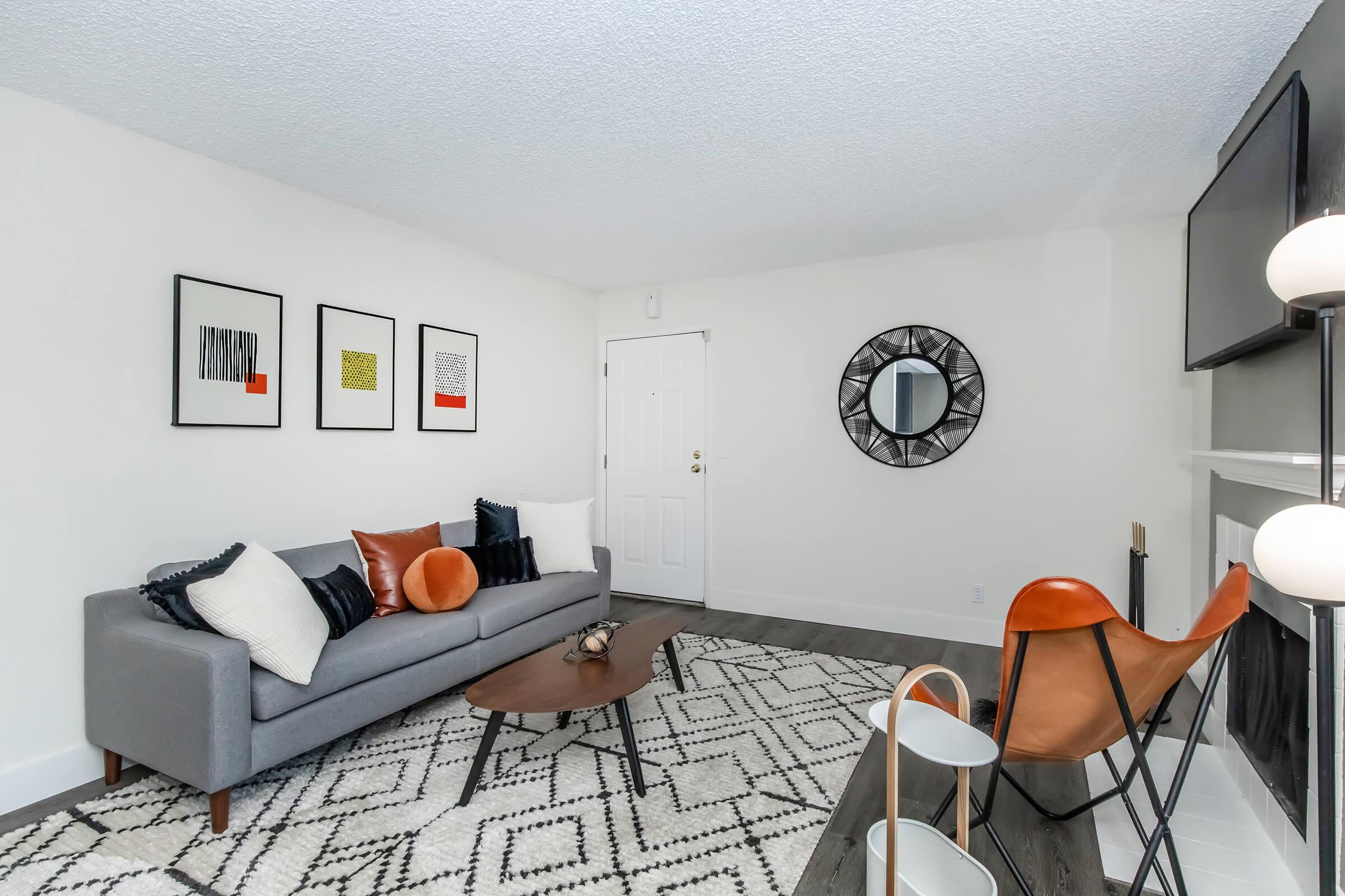 A modern living room featuring a gray sofa with decorative pillows, a round wooden coffee table, and a patterned area rug. There are framed artworks on the wall, a circular mirror, and a stylish chair. The space has light walls and a television mounted above a minimalistic fireplace.