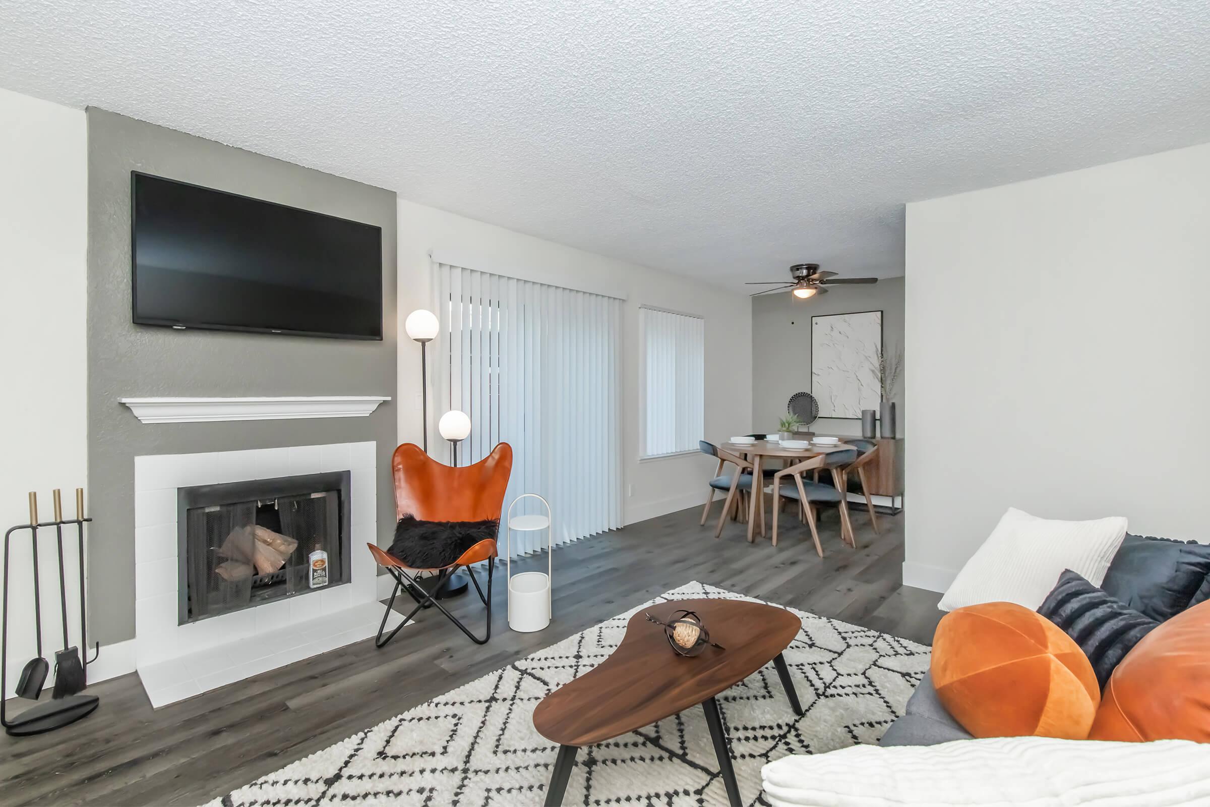 A modern living room featuring a stylish fireplace with a TV above, a cozy brown leather chair, a black sofa with decorative pillows, and a geometric area rug. A dining area with a wooden table and chairs is visible in the background, complemented by large windows with blinds for natural light.