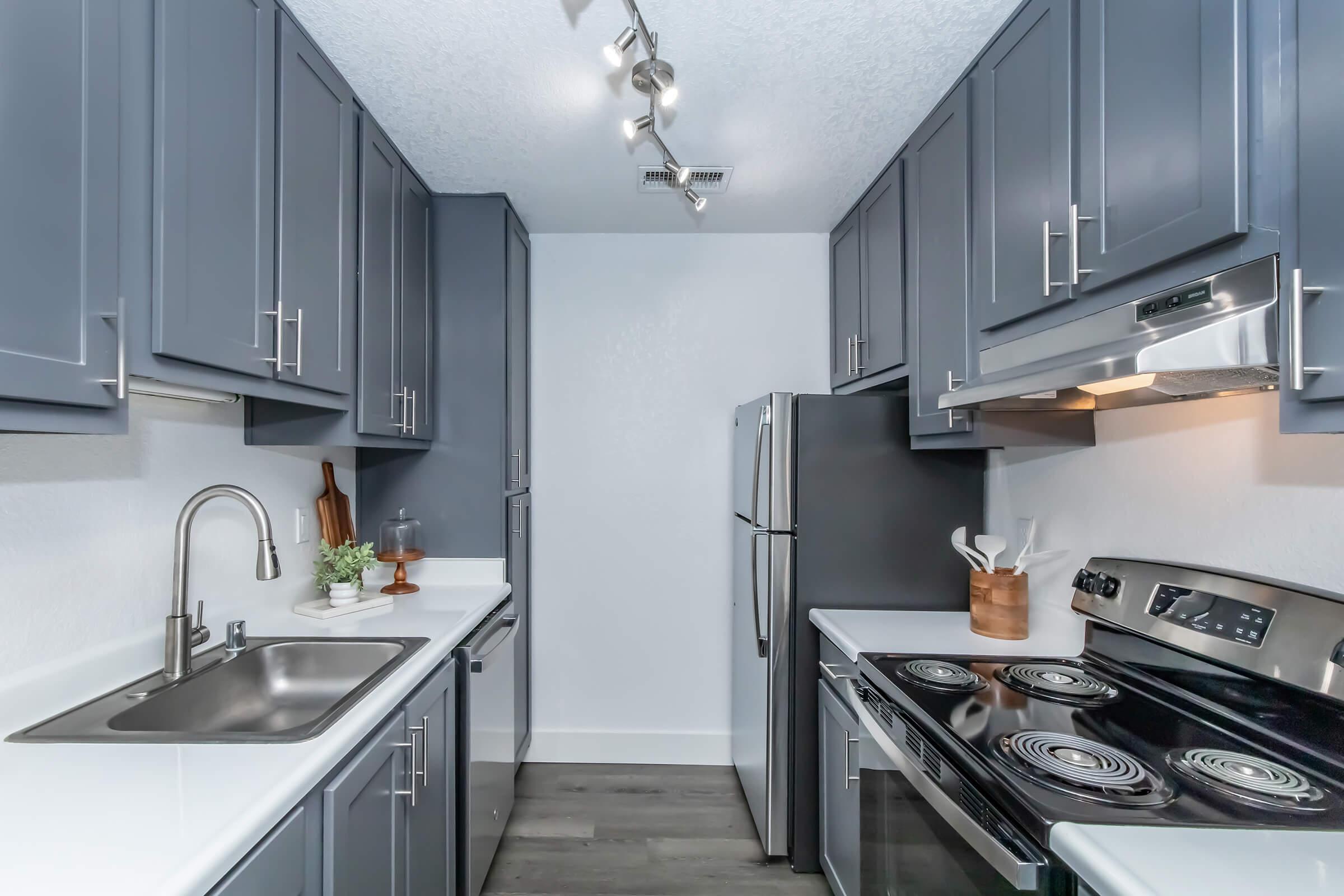 Modern kitchen with gray cabinets and a white countertop. Features a stainless steel refrigerator, oven, and sink. Bright lighting from a track light above. The design is functional and contemporary, with a clean, minimalist aesthetic.