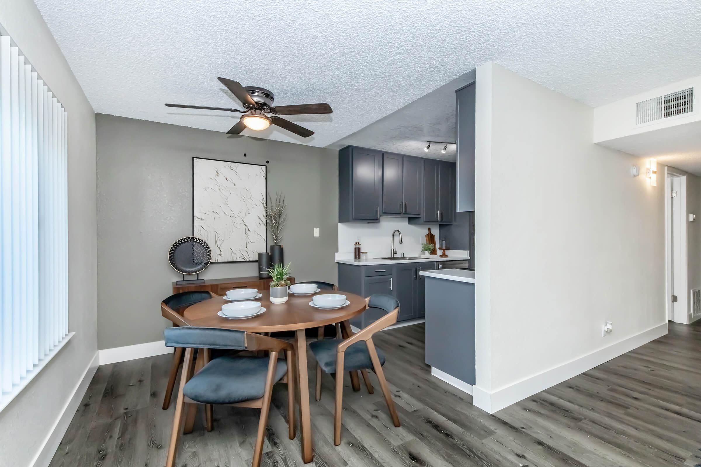 A modern dining area featuring a round wooden table set for four with blue chairs. In the background, a gray kitchen is partially visible, with dark cabinets and a small counter. A ceiling fan is mounted above, and a minimalist wall art piece is displayed beside the table. The flooring is a sleek, dark laminate.