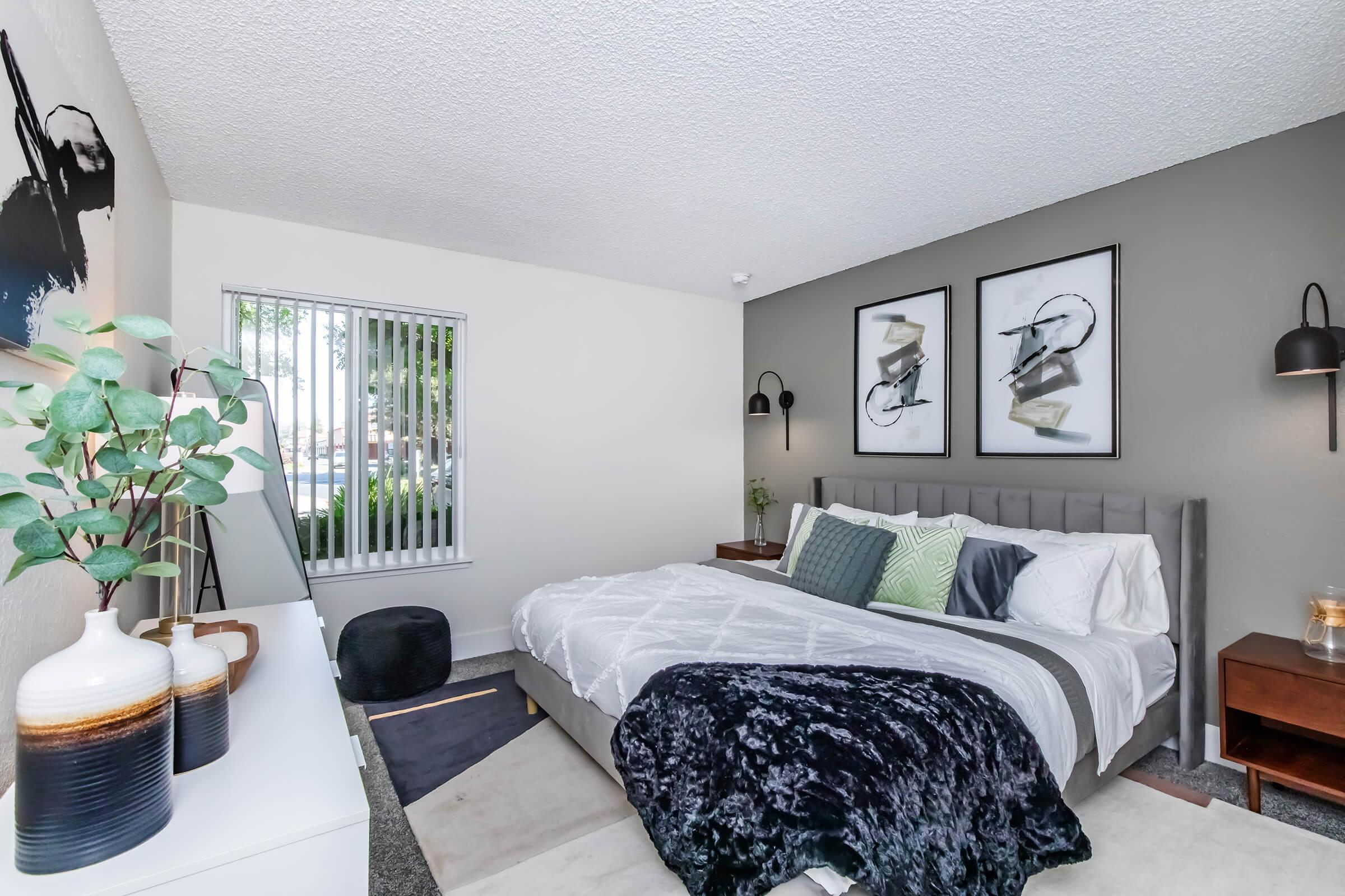 A modern bedroom featuring a gray upholstered bed with decorative pillows, a cozy black throw blanket, and two framed artworks on the wall. There are bedside tables with lamps, a white dresser, and a potted plant. Natural light filters through window blinds, illuminating the space.