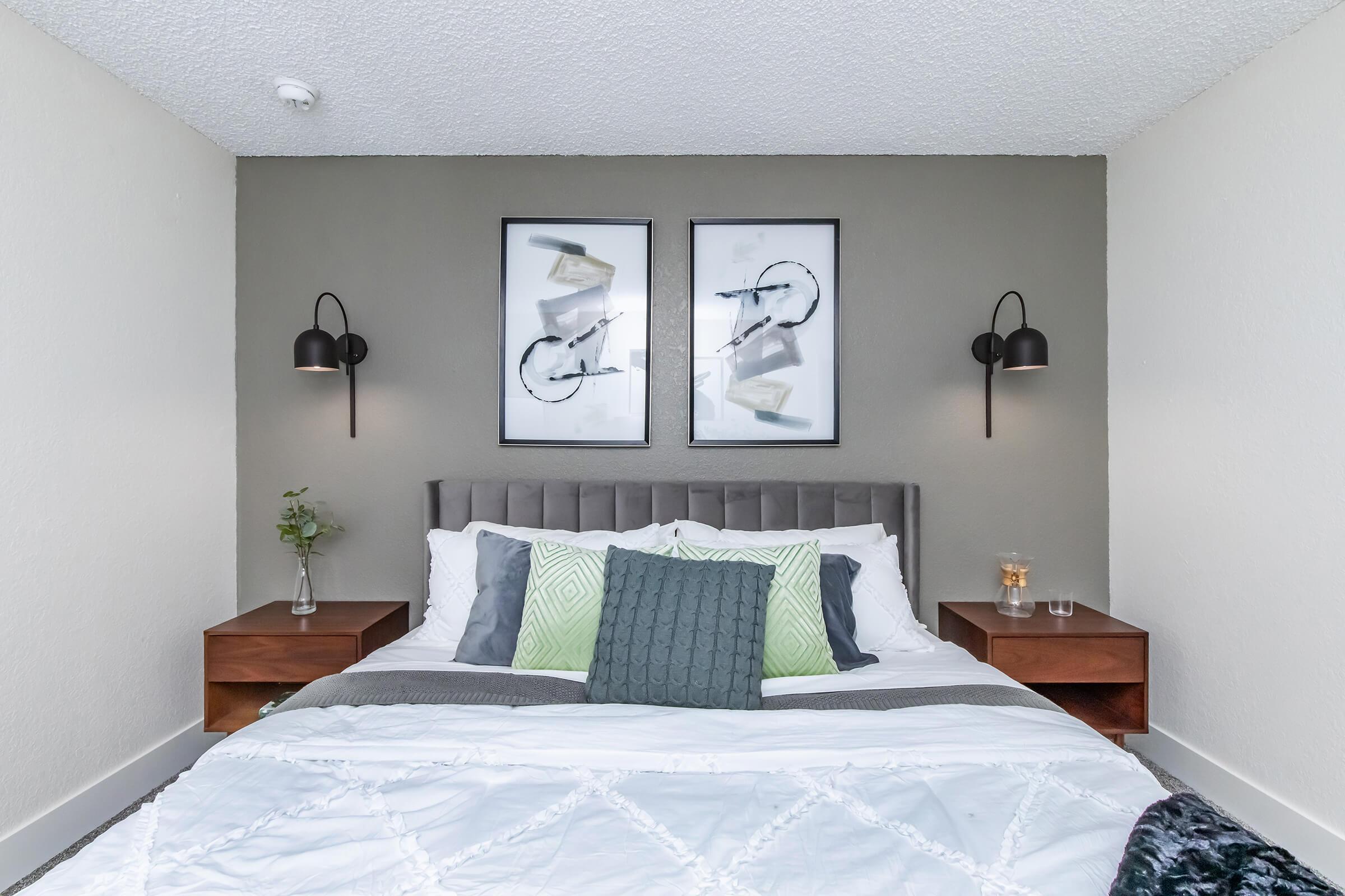 A modern bedroom featuring a gray upholstered headboard with decorative pillows, framed artwork on the wall, and stylish wall sconces. There are wooden nightstands on either side of the bed, one with a decorative plant and a glass. The walls are light-colored, creating a cozy ambiance.
