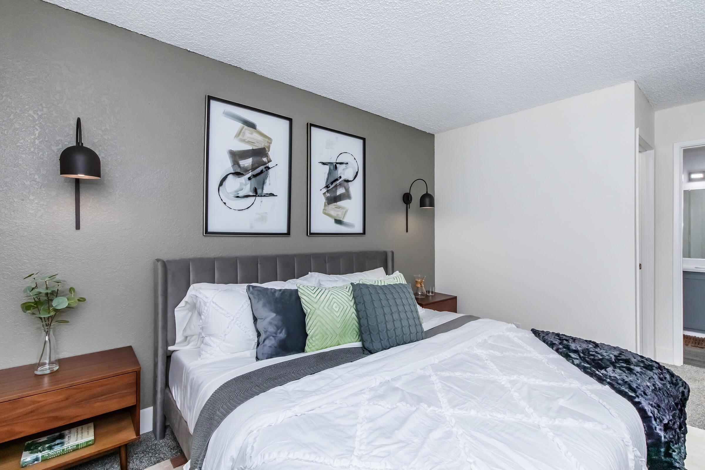 A cozy bedroom featuring a neatly made bed with decorative pillows and a stylish gray headboard. On the walls, there are two framed prints of bicycles. Soft lighting from wall sconces complements the modern furniture, which includes a wooden nightstand with a small plant. The room is designed with neutral colors for a calming atmosphere.