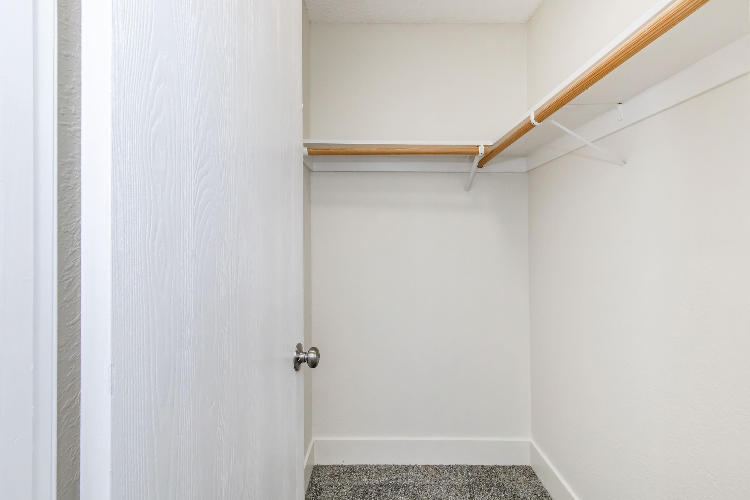 An empty closet with a white door and a simple wooden hanging rod. The walls are painted a light color, and the floor is covered with a grey textured surface. The space is well-lit, highlighting the clean and minimalistic design.
