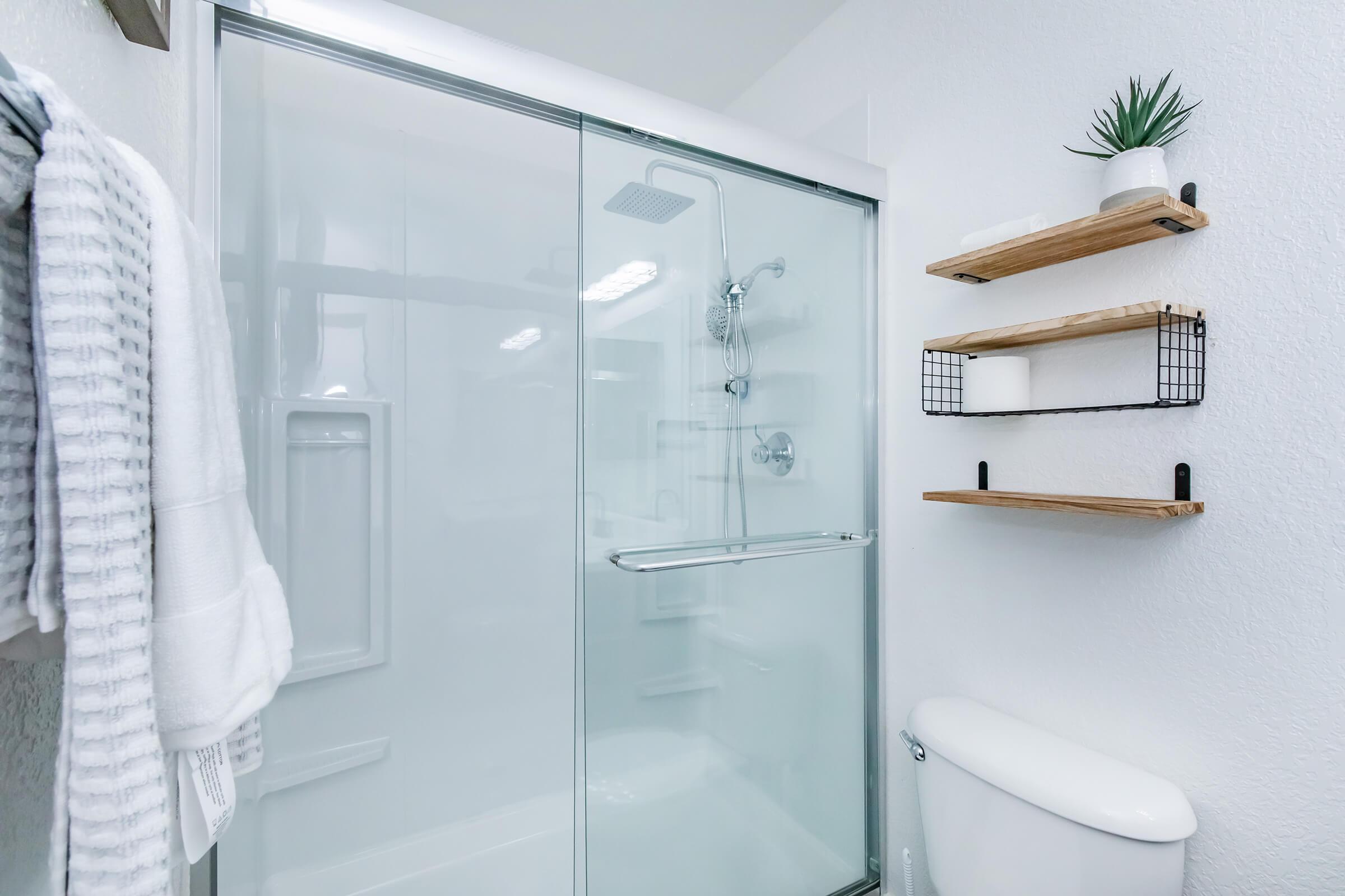 A modern bathroom featuring a glass shower enclosure, a white toilet, and wooden shelves mounted on the wall. The shelves hold toiletries and a small potted plant. A towel hangs neatly on a hook, contributing to a clean and contemporary aesthetic. Soft lighting enhances the bright, airy atmosphere.