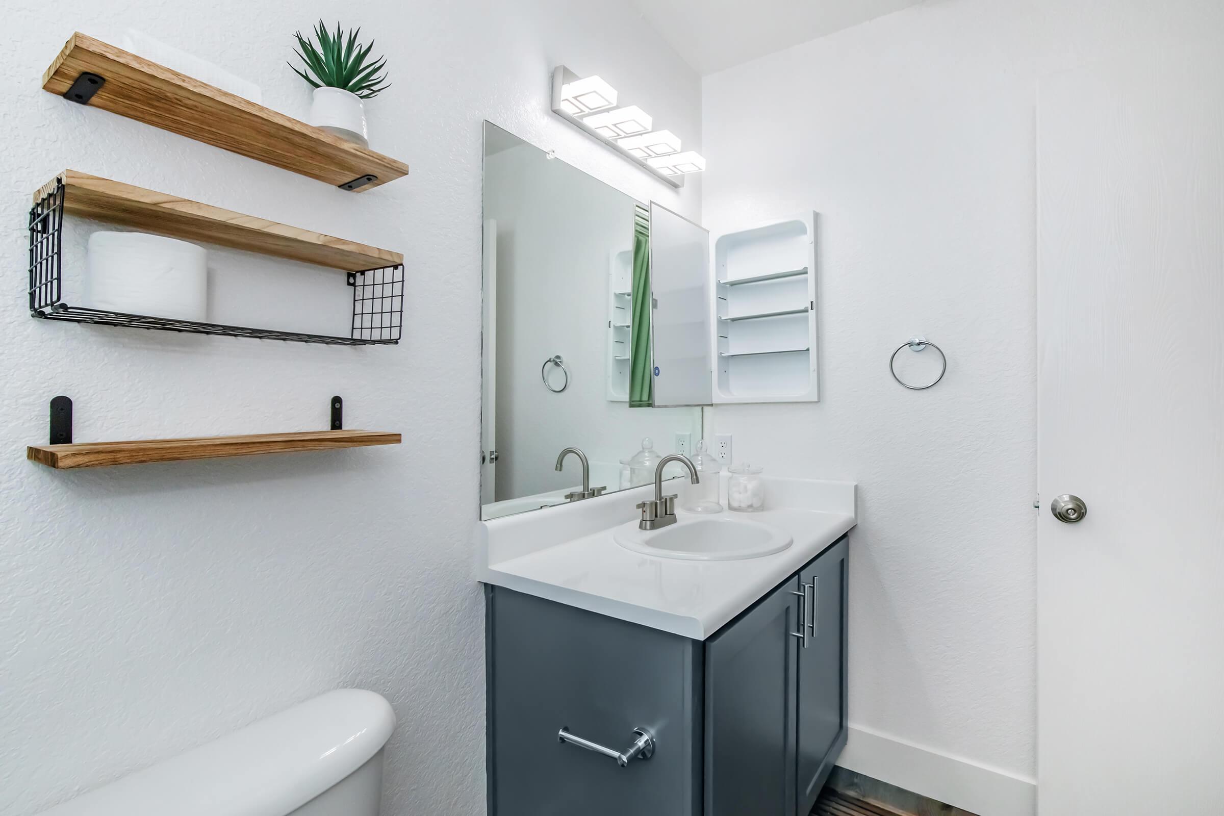 A modern bathroom featuring a white wall, gray vanity with a sink, and a large mirror. There are wooden shelves displaying a small plant and toiletries. A white towel is hung on a silver towel ring. A white toilet is in the corner, and a door is visible on the right side.