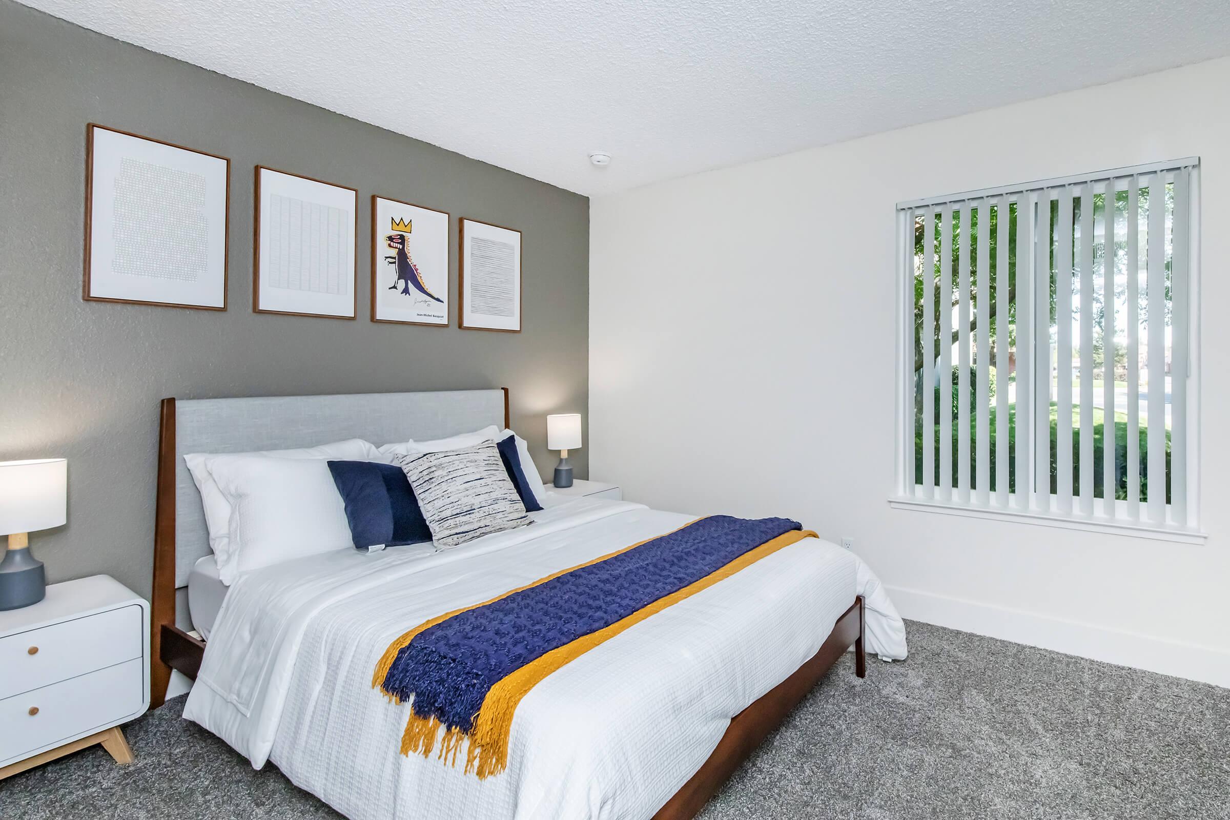 Cozy bedroom featuring a comfortable bed with white linens and a blue throw blanket. The room has stylish wall art above the bed, bedside lamps on sleek nightstands, and a window with vertical blinds allowing natural light in. The carpeted floor adds to the warm ambiance of the space.