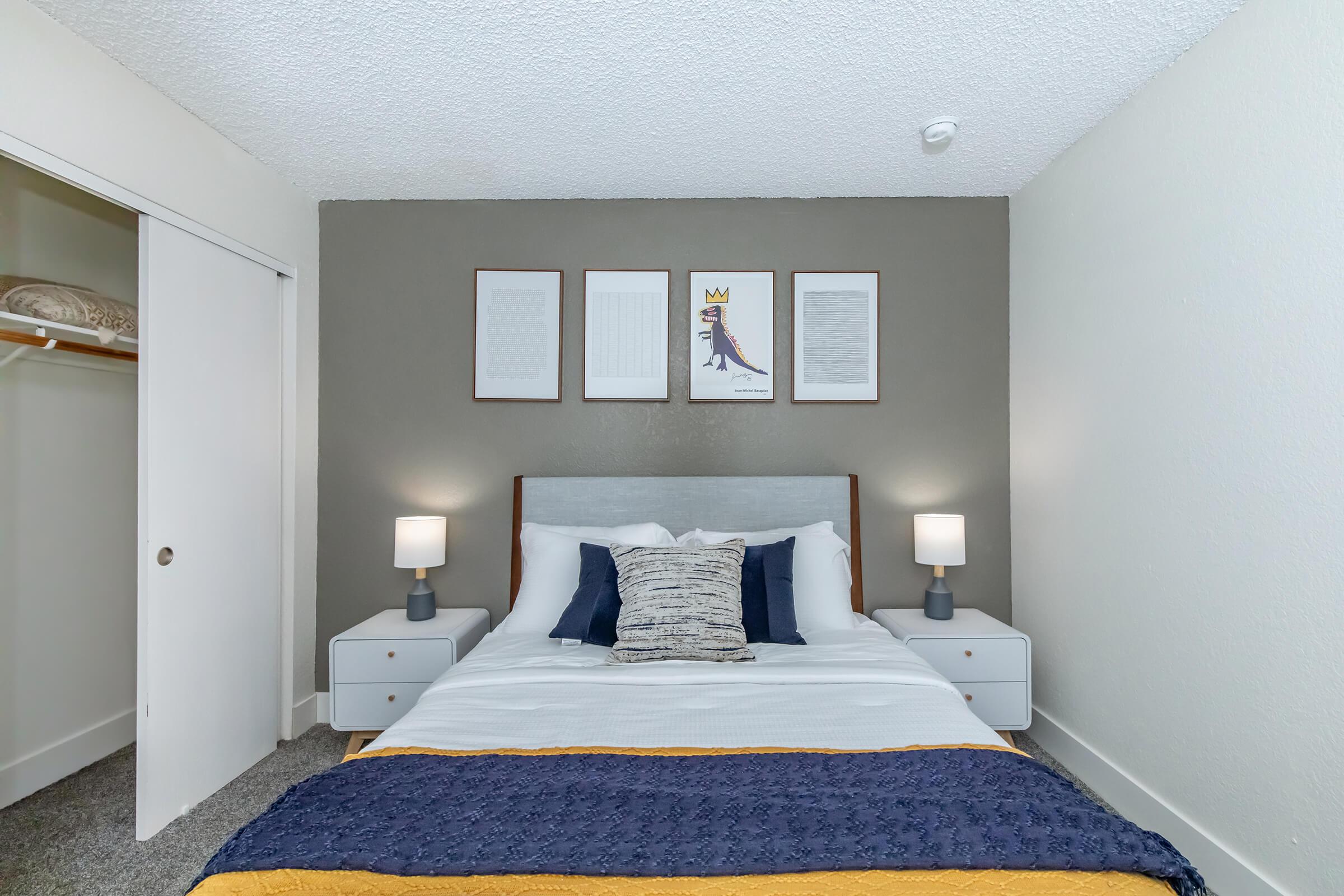 A cozy bedroom featuring a double bed with white bedding and decorative pillows, flanked by two nightstands with lamps. The wall behind the bed is painted gray and displays four framed artworks. A closet is visible to the left, and the floor is covered with gray carpet. The overall ambiance is welcoming and modern.