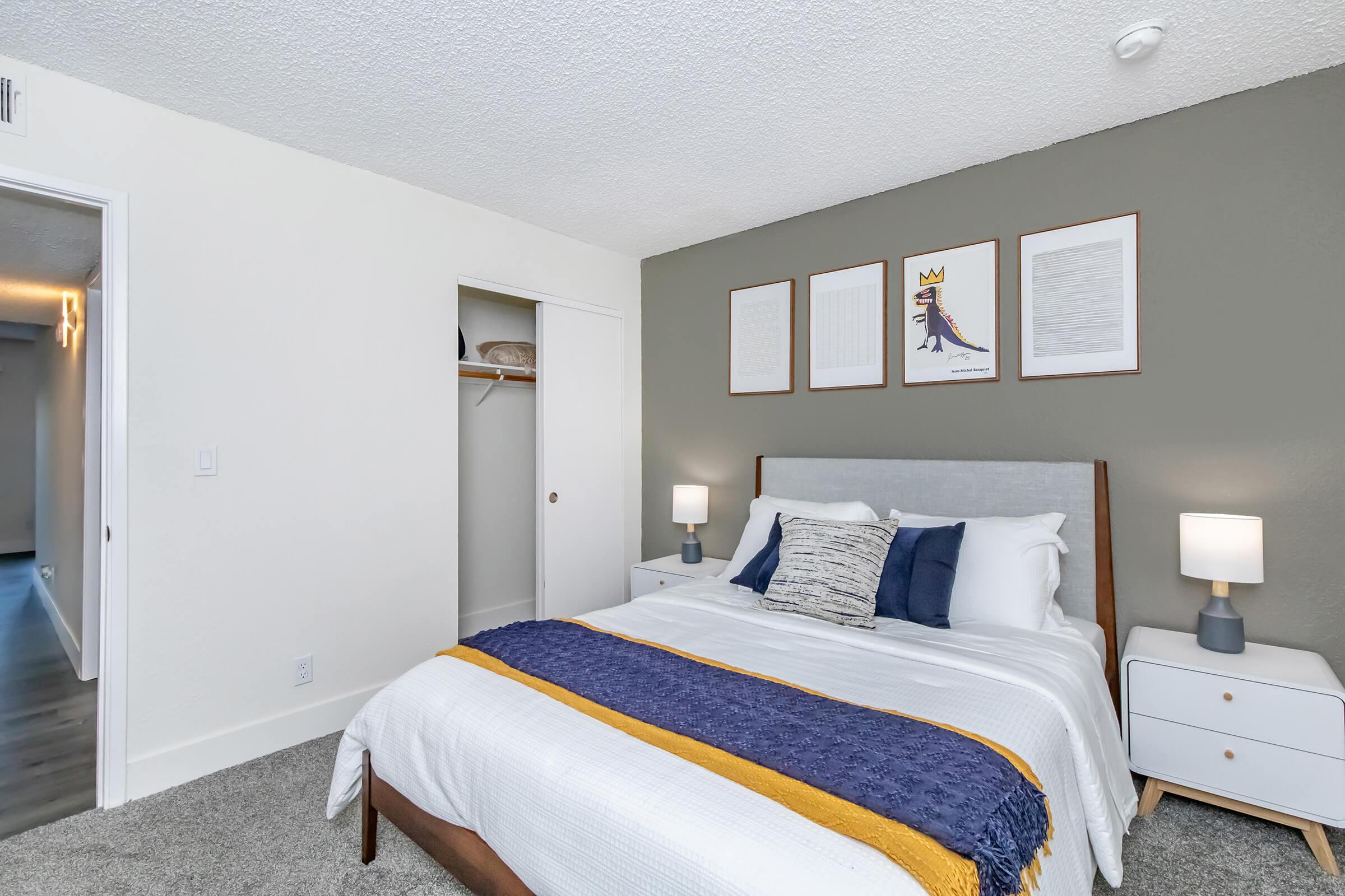 A cozy bedroom featuring a neatly made bed with a gray headboard, white bedding, and decorative pillows. There are two bedside lamps on white nightstands. A wall with three framed art posters and a dark accent wall. A closet is visible in the corner, and a doorway leads to another room.