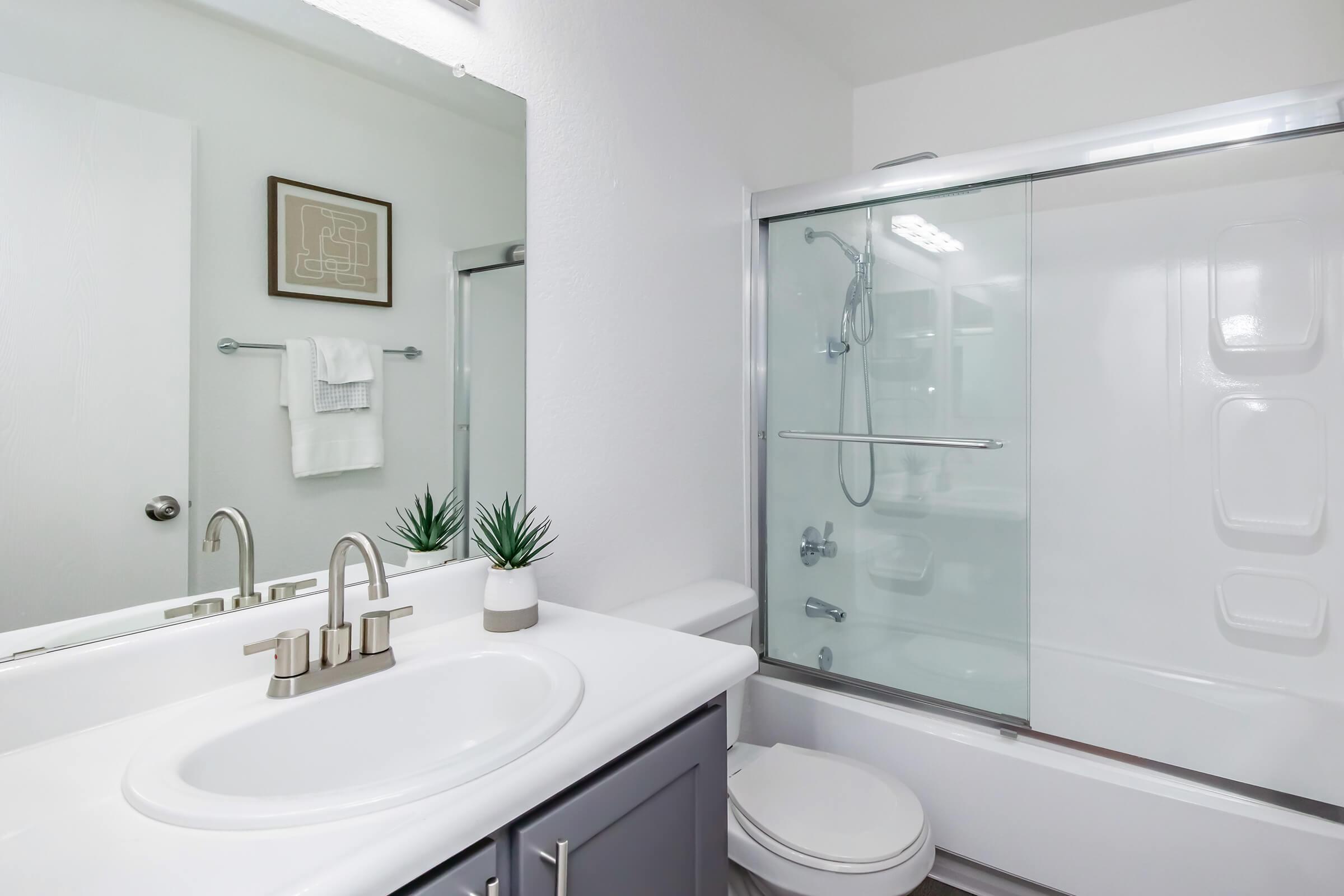 A modern bathroom featuring a white sink with a stylish faucet, a large mirror, and a shower with a glass door. There is a small plant on the counter and neatly arranged towels on a rack. The walls are painted in a light color, creating a bright and clean atmosphere.