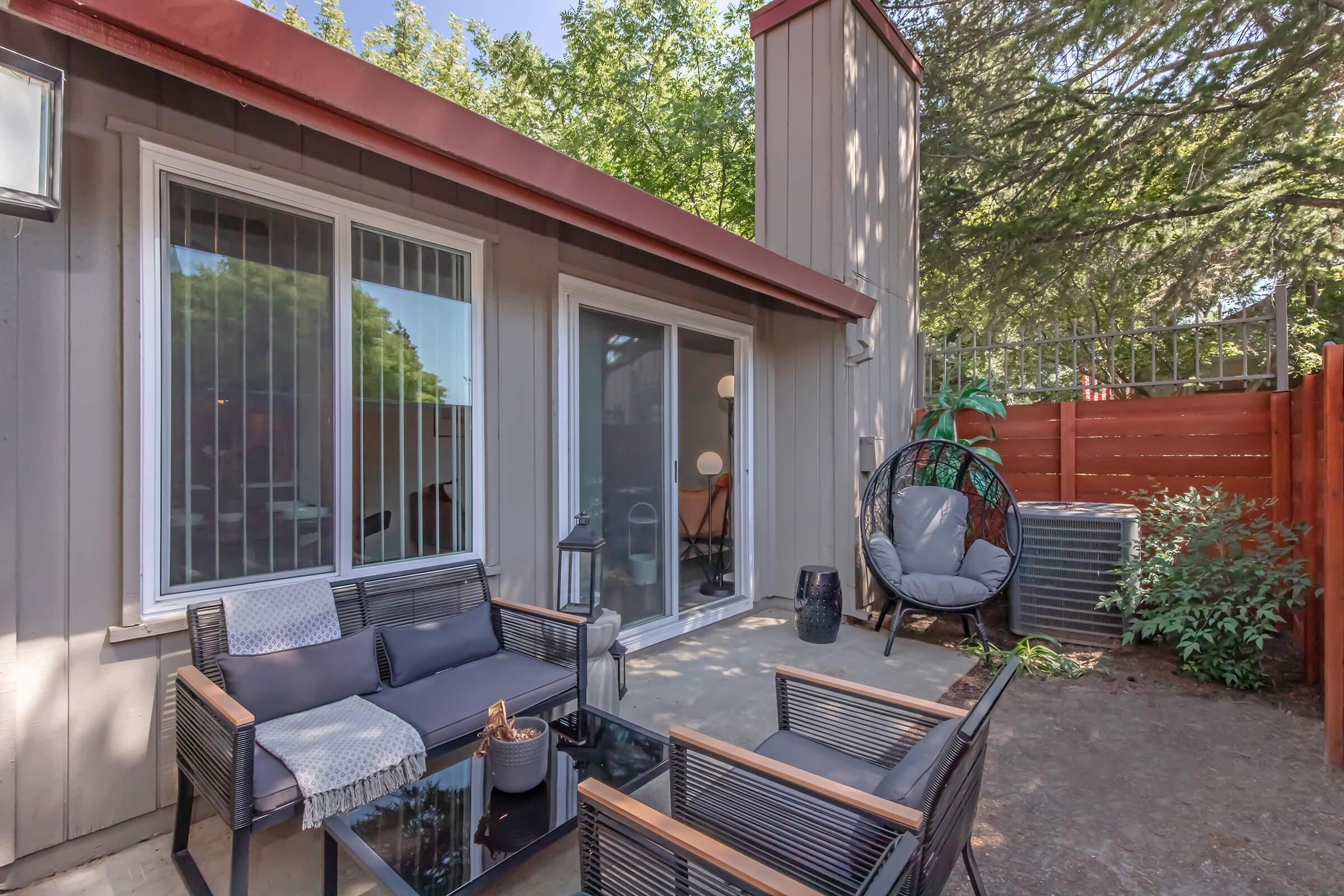 Cozy outdoor patio area featuring modern seating, a glass table, and a hanging chair. Surrounded by trees and a wooden fence, it creates a tranquil space for relaxation. Large sliding glass doors provide access to an interior room, enhancing the inviting atmosphere.