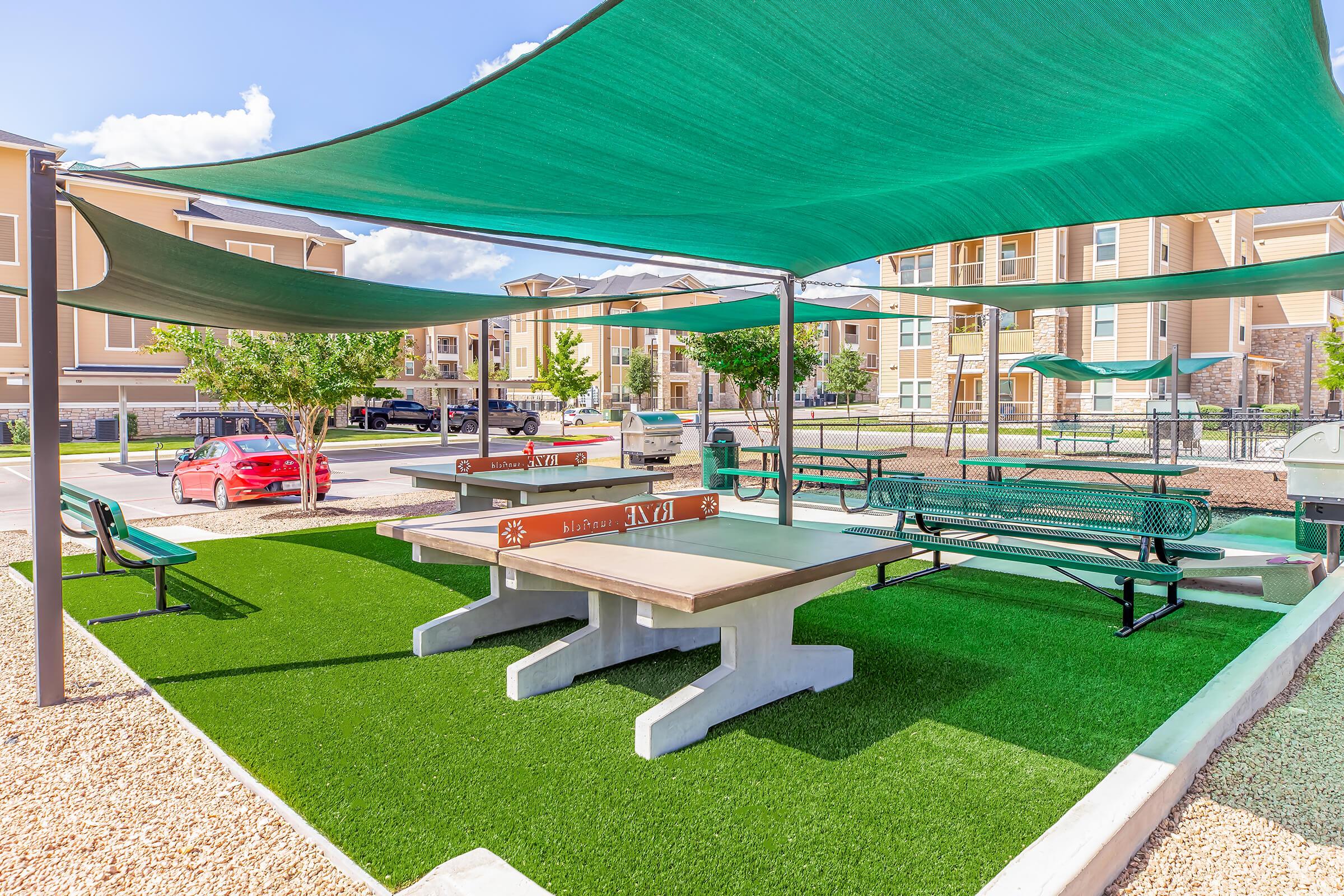 Outdoor recreational area featuring shaded picnic tables, green artificial grass, and benches. Surrounding residential buildings are visible in the background, along with a parked red car. The scene is bright and open, ideal for social gatherings or relaxation.