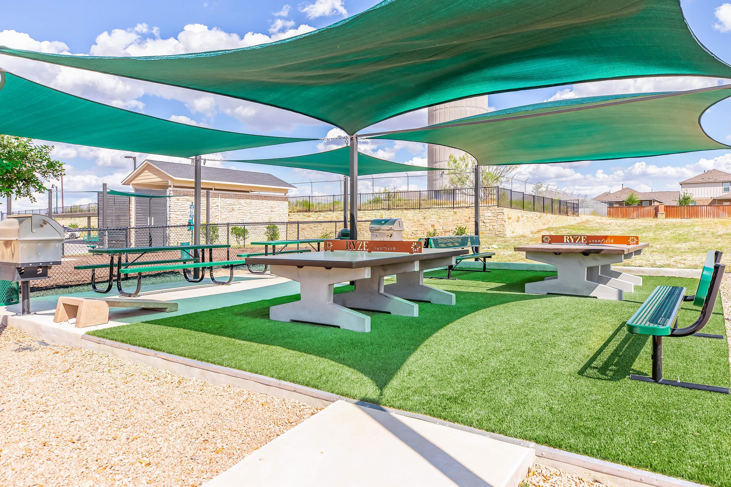A recreational area featuring shaded picnic tables and benches on artificial grass. There are two concrete tables for games, a grill area nearby, and a fenced background with a few structures. Bright blue sky with clouds above, creating a spacious outdoor atmosphere.