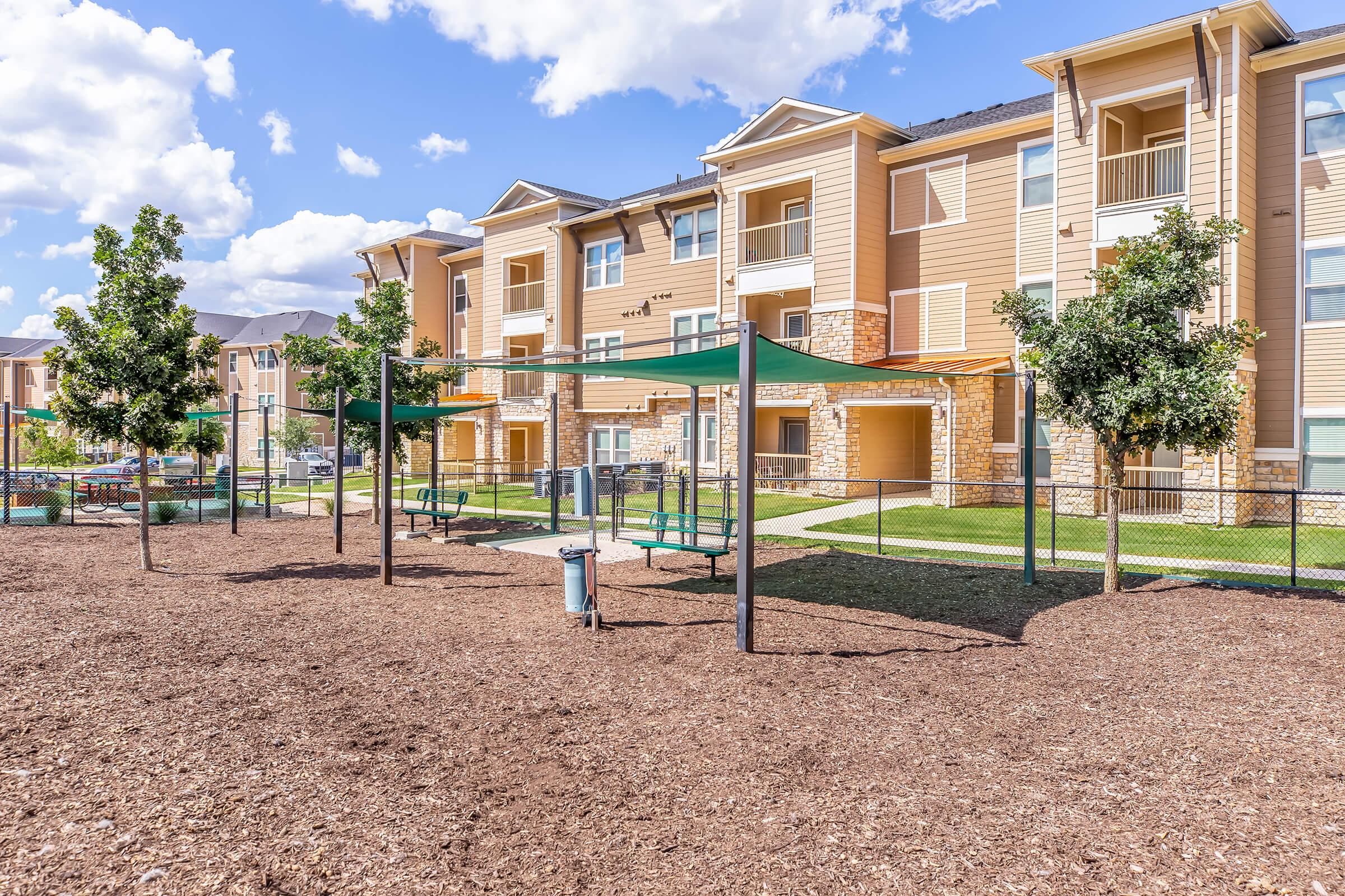 A modern apartment complex with multiple buildings, featuring a playground area with green shade structures, benches, and mulch landscaping. Clear blue sky with fluffy clouds above.