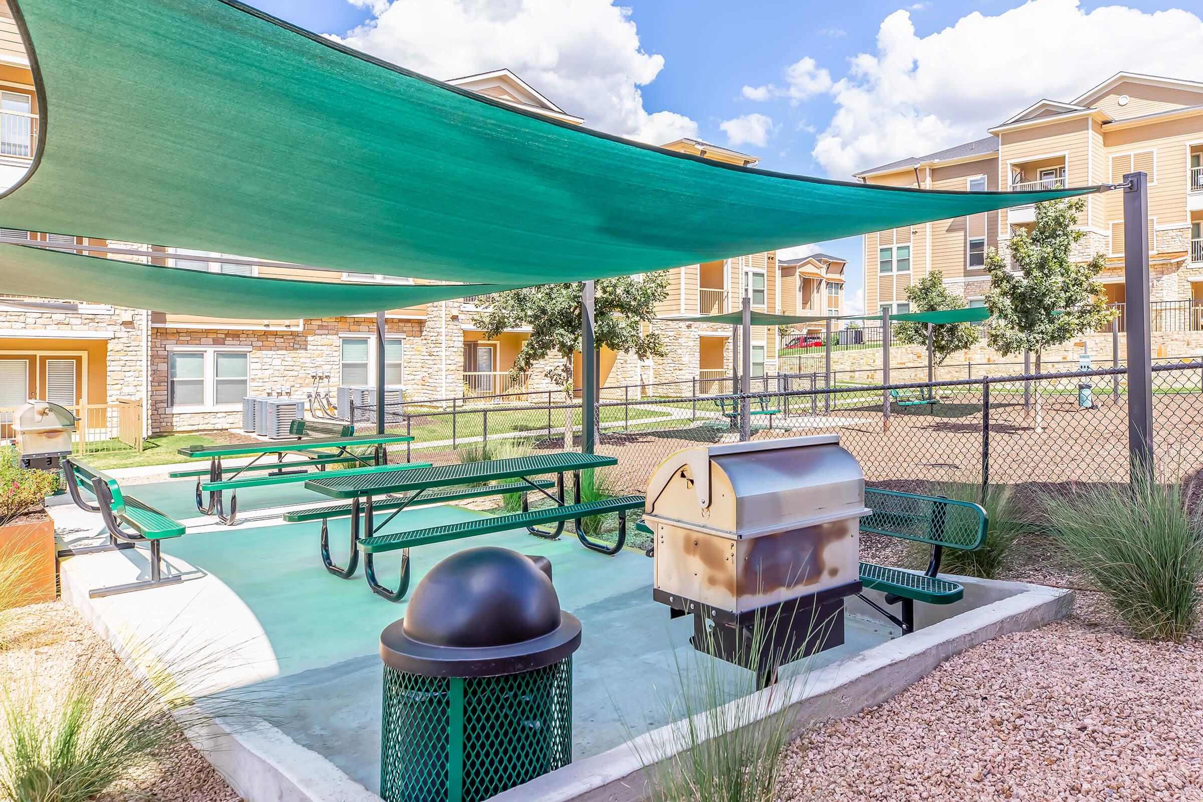 Outdoor barbecue area with picnic tables under green shade structures, surrounded by landscaped gravel and grass. An apartment complex is visible in the background, featuring multiple buildings and trees. A trash bin is also present nearby.