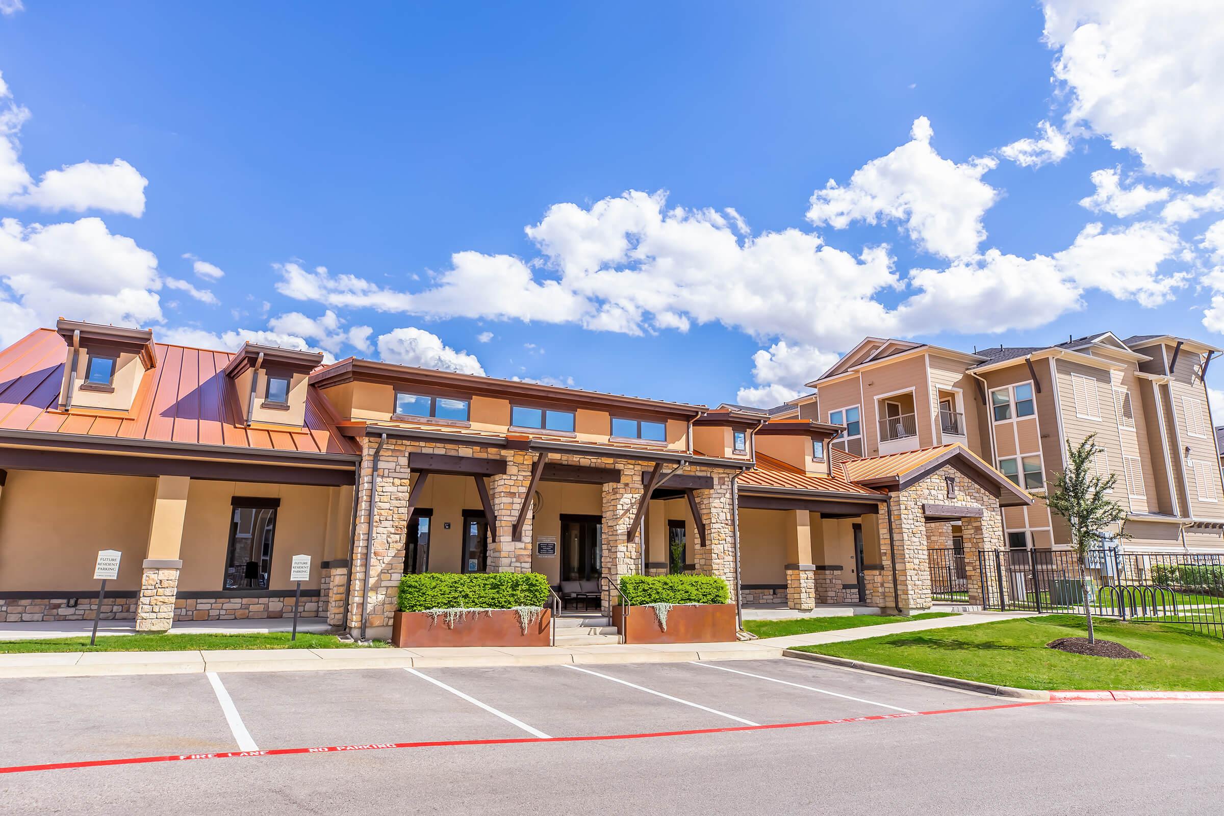 A well-lit, modern building with a stone and metal facade, featuring a covered entrance and large windows. The parking lot in front has several empty spaces. In the background, there is a larger residential building, and the sky is partly cloudy with blue patches.