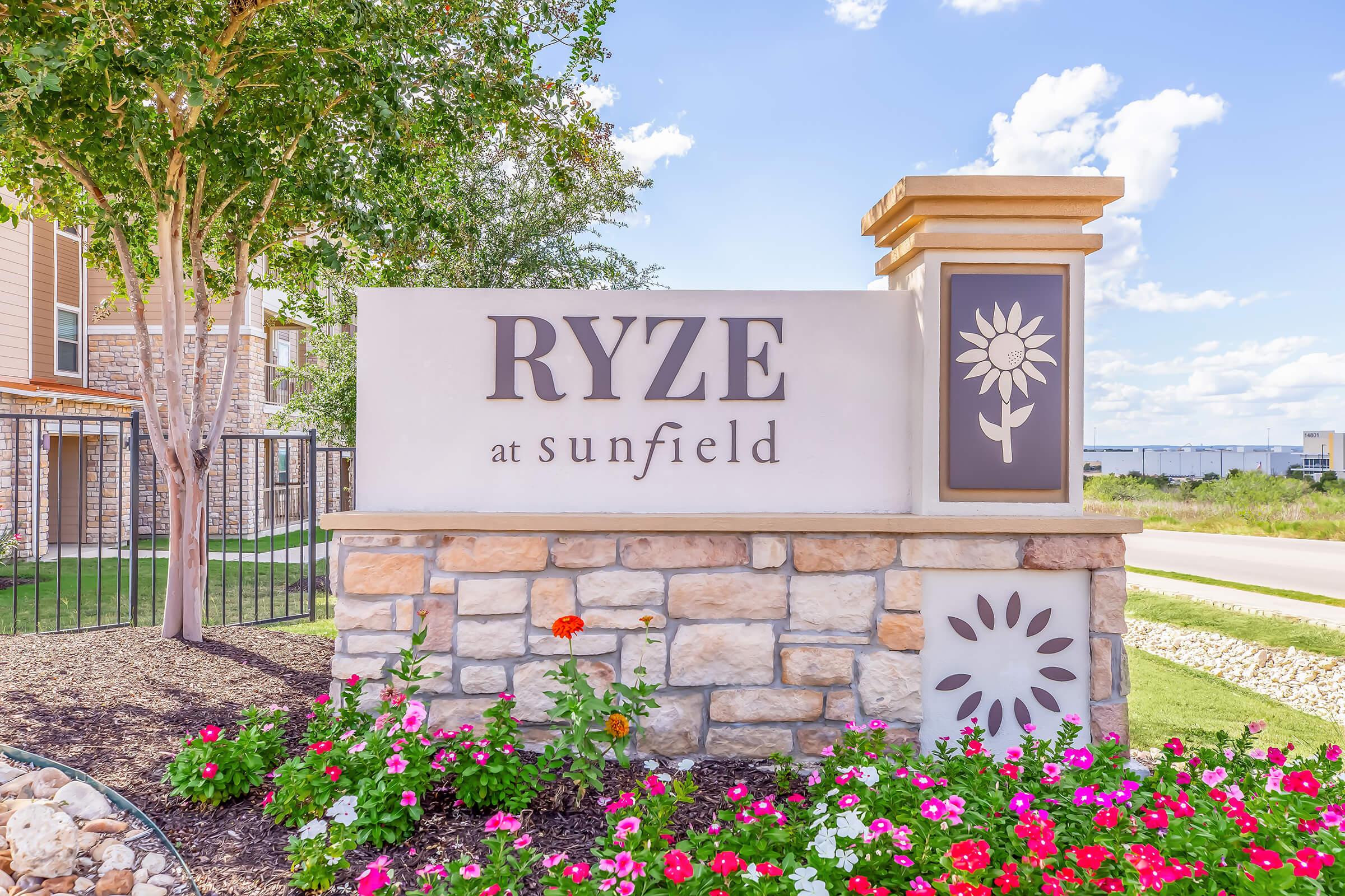 Sign for "RYZE at Sunfield" featuring a decorative stone base and a floral design. Surrounded by colorful flowers and green landscaping, with a blue sky and fluffy clouds in the background.