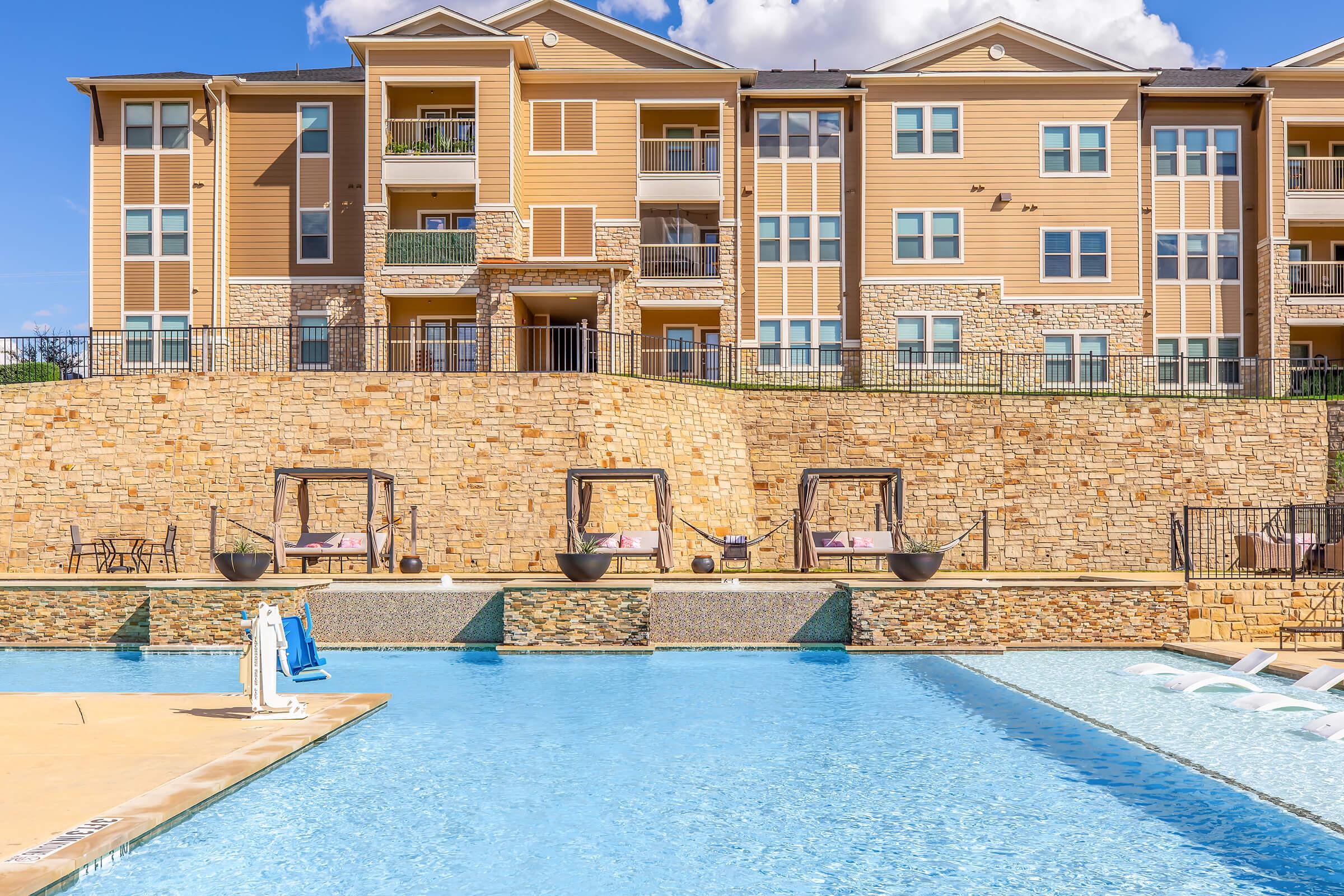 A clear blue swimming pool in the foreground, with a stone wall and several hanging lounge chairs positioned beside it. In the background, there are three-story tan apartment buildings with balconies and large windows under a bright blue sky with scattered clouds.